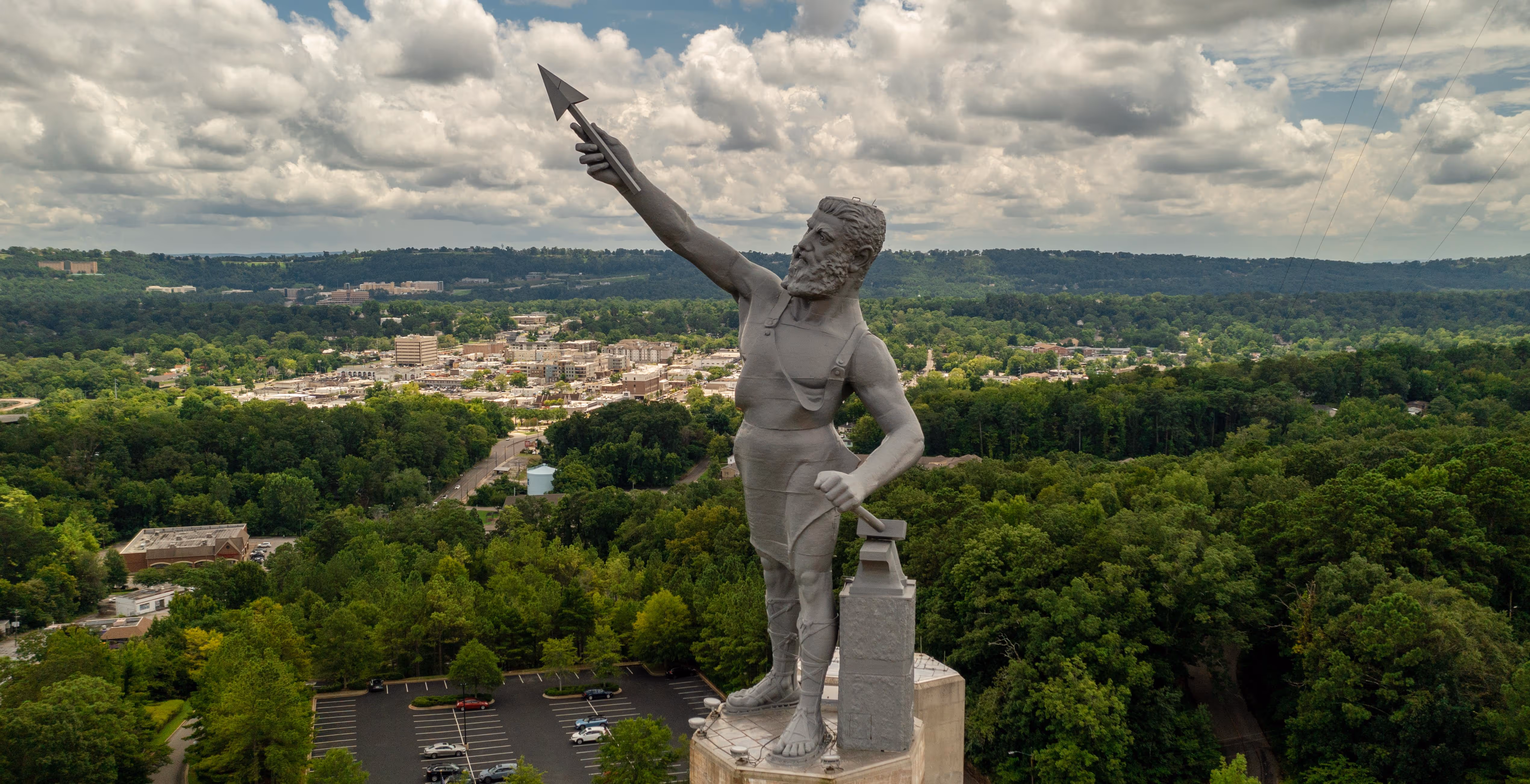 The Vulcan statue, a large iron statue of a bearded man holding an arrow, overlooking a cityscape surrounded by green trees under a cloudy sky.