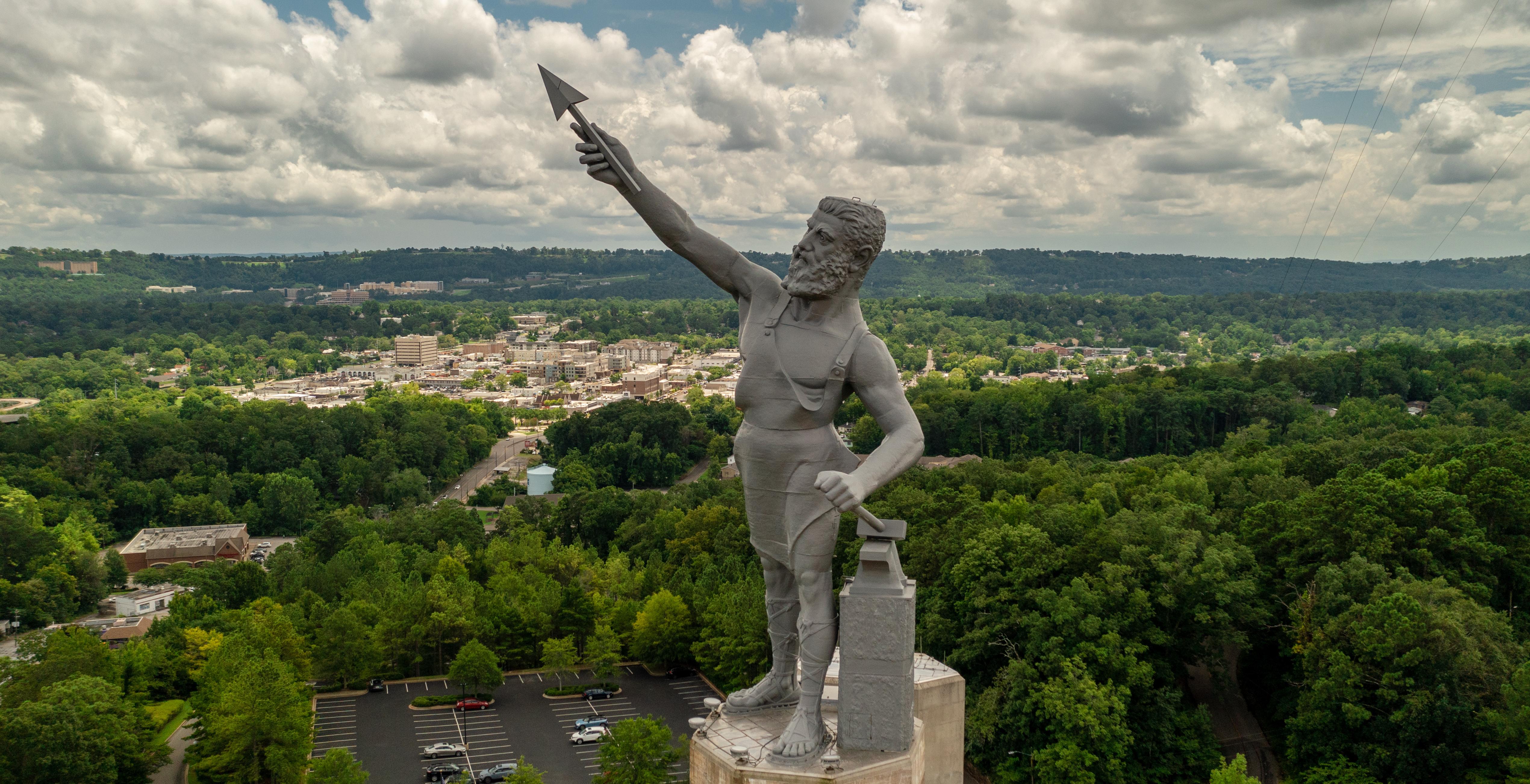The Vulcan statue, a large iron statue of a bearded man holding an arrow, overlooking a cityscape surrounded by green trees under a cloudy sky.