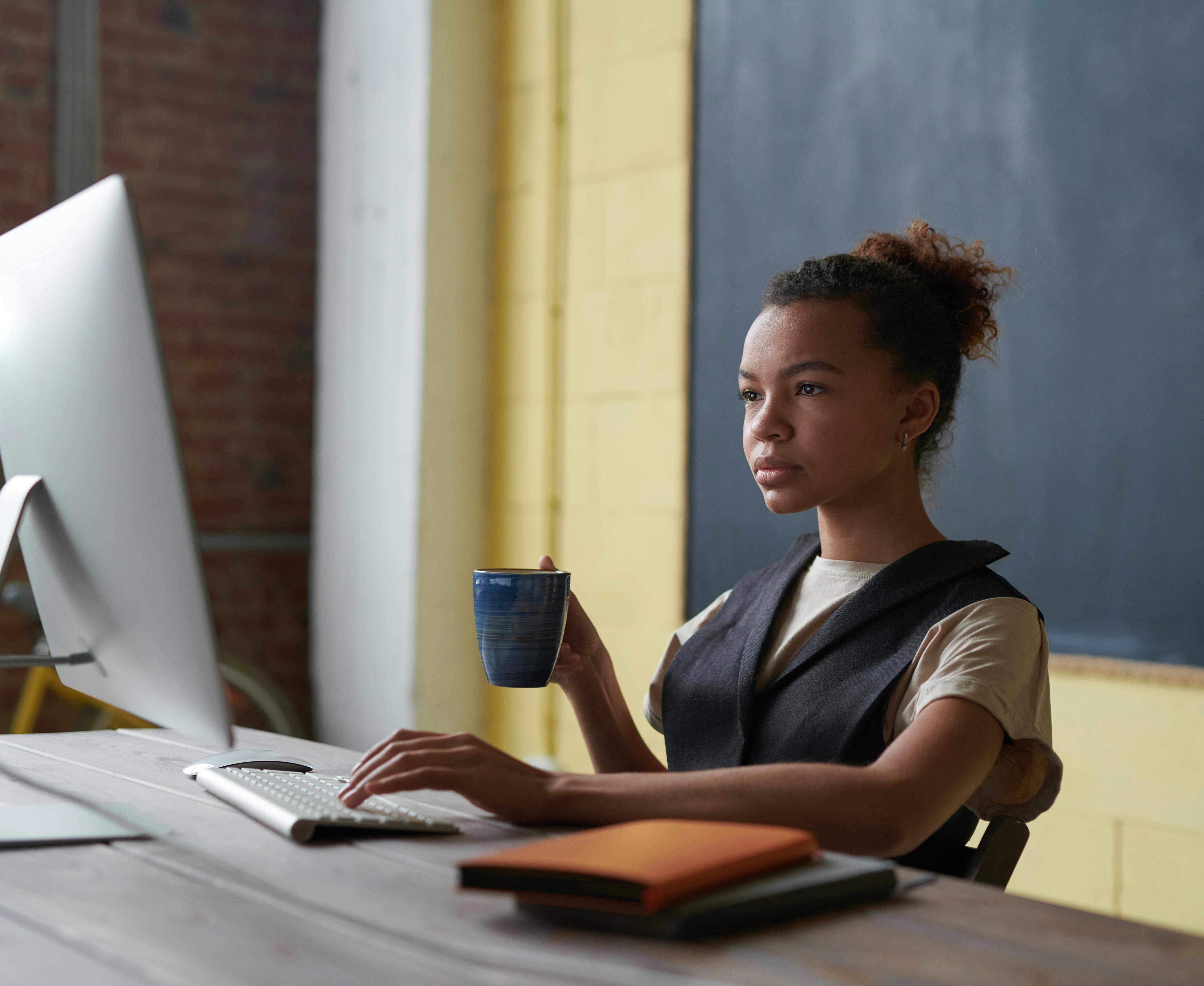 Vrouw die achter een bureau zit, met een blauw kopje in haar hand en kijkt naar een computerbeeldscherm.