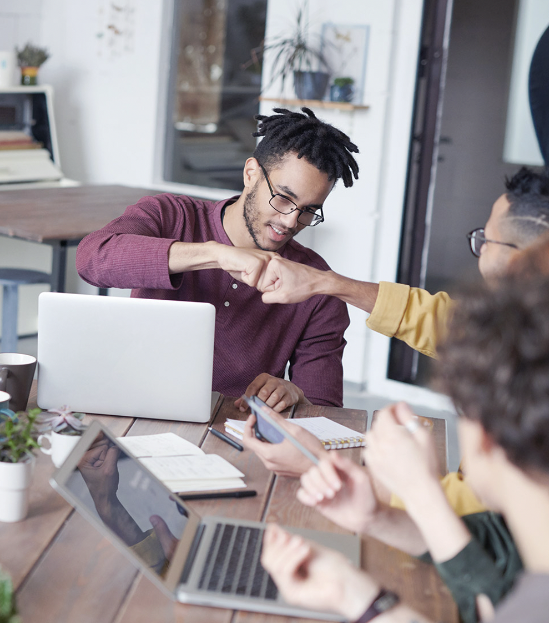 Twee mannen geven elkaar een boks terwijl ze aan een tafel met laptops en notitieboekjes zitten.