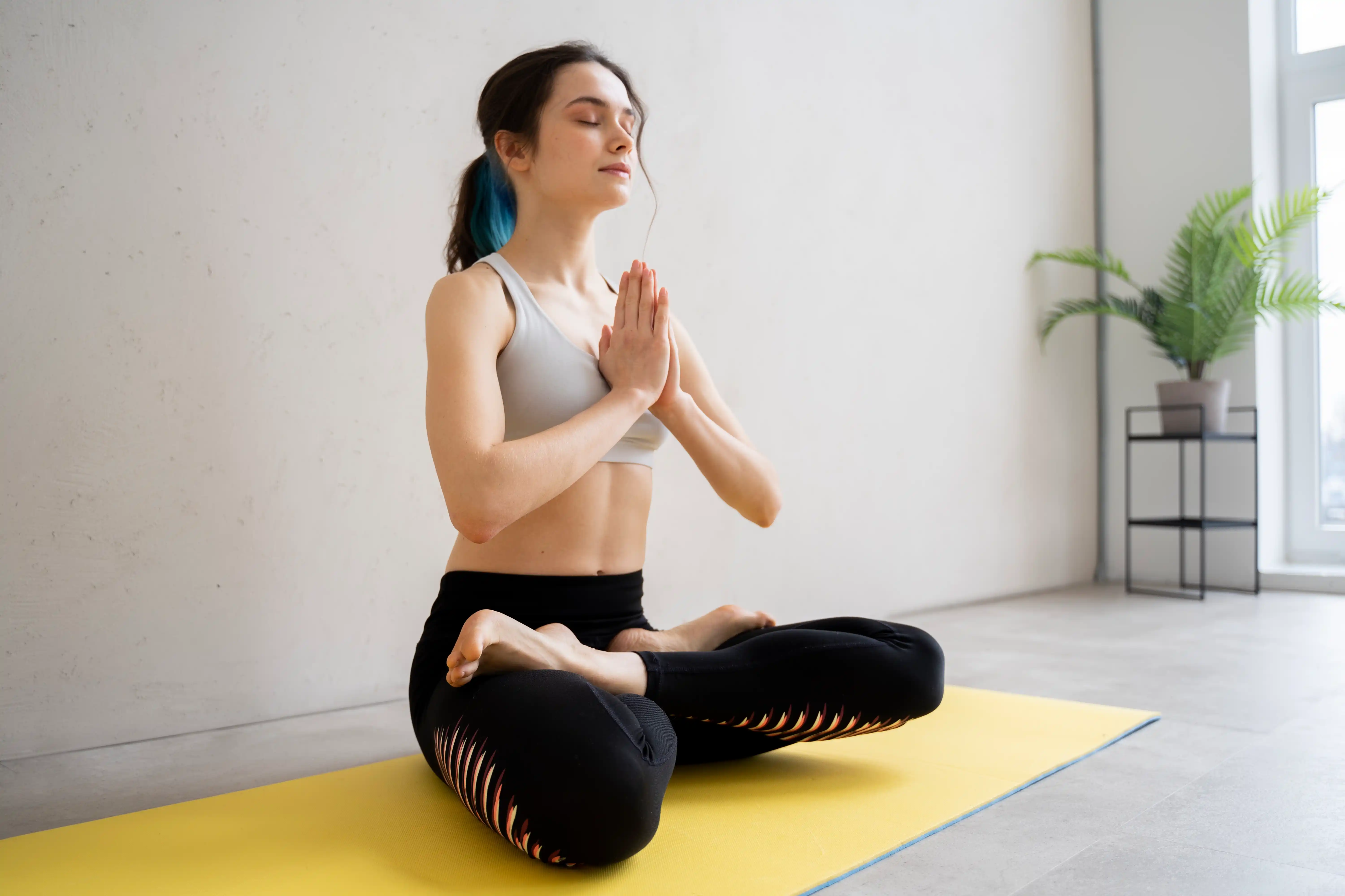 Young woman with blue-tipped hair meditating in lotus pose on a yellow mat in a bright, minimalist room.