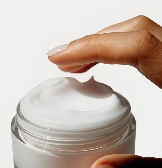 Hand with manicured nails dipping finger into white cream in an open jar.