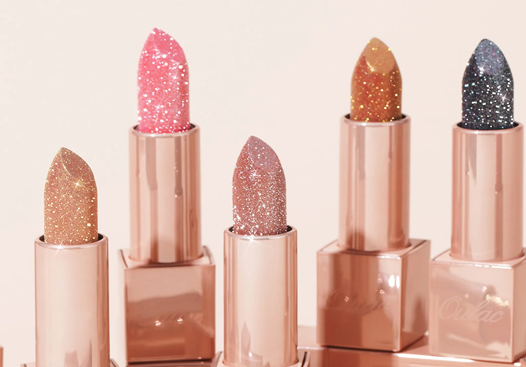 Five glittery lipsticks in rose gold tubes arranged in a row with colors ranging from gold to pink and dark purple.