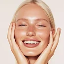 Smiling blonde woman with glowing skin gently touching her face with both hands.