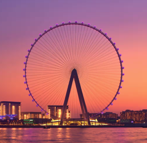 The illuminated Dubai Eye ferris wheel at sunset with surrounding buildings and water in the foreground.