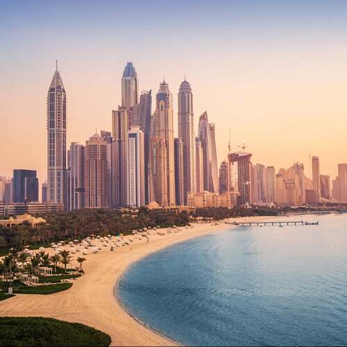 Dubai Marina skyline with tall skyscrapers along the coastline and a sandy beach at sunset.