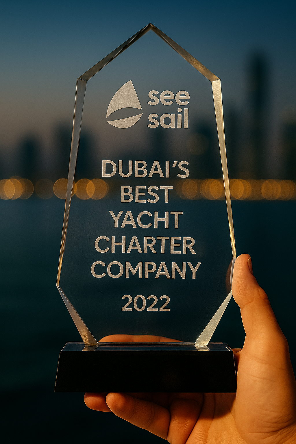 Hand holding a transparent crystal award labeled 'see sail DUBAI'S BEST YACHT CHARTER COMPANY 2022' against a blurred cityscape at dusk.