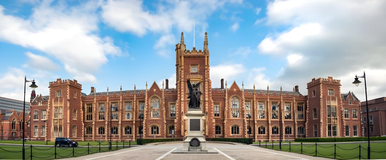 Historic red brick building with large windows, a central clock tower, and a statue in the foreground under a partly cloudy blue sky.