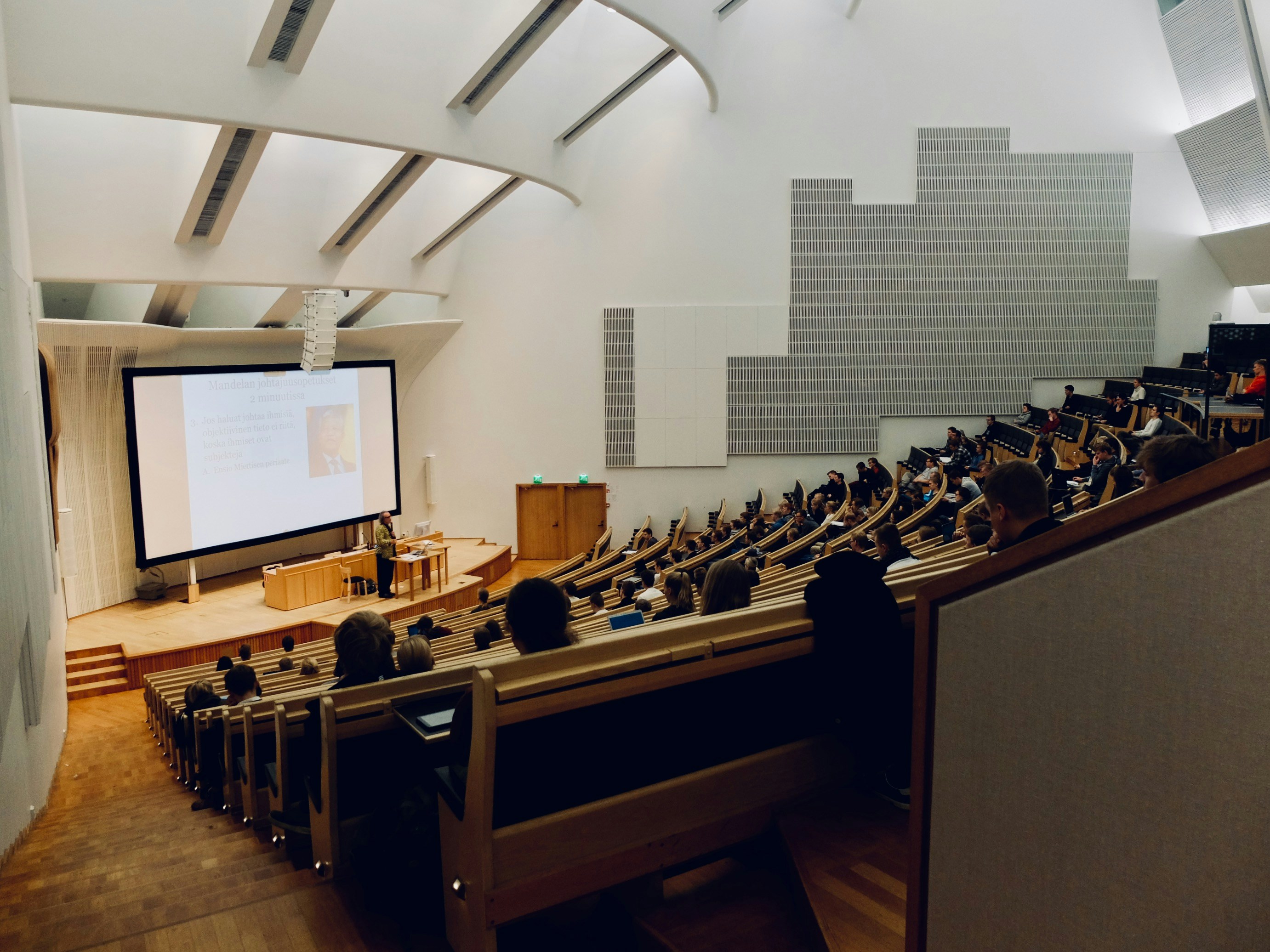 Audience seated in a large lecture hall watching a speaker presenting slides on a large screen.