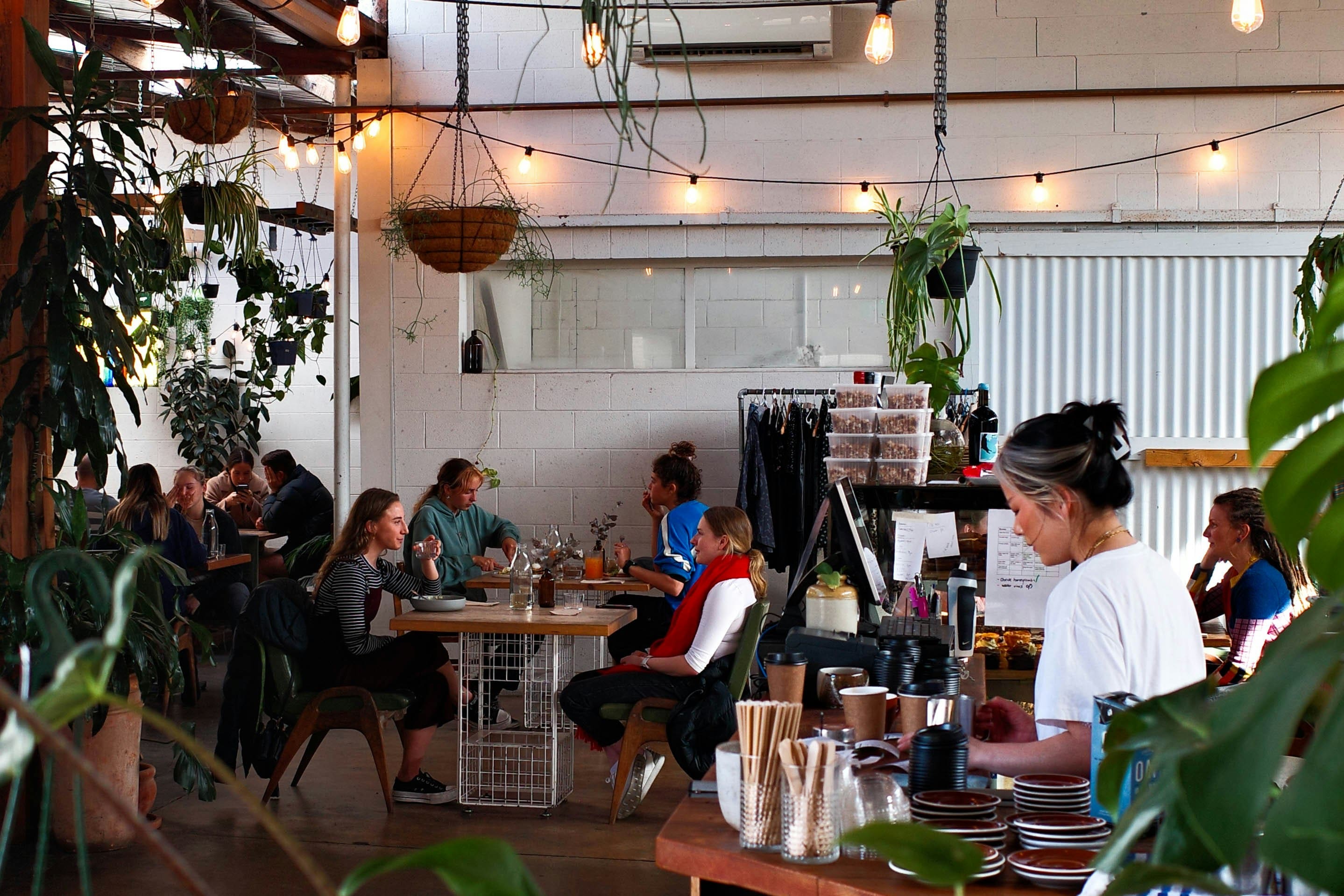 Busy café interior with people sitting at tables chatting and a barista working behind the counter surrounded by plants and warm string lights.