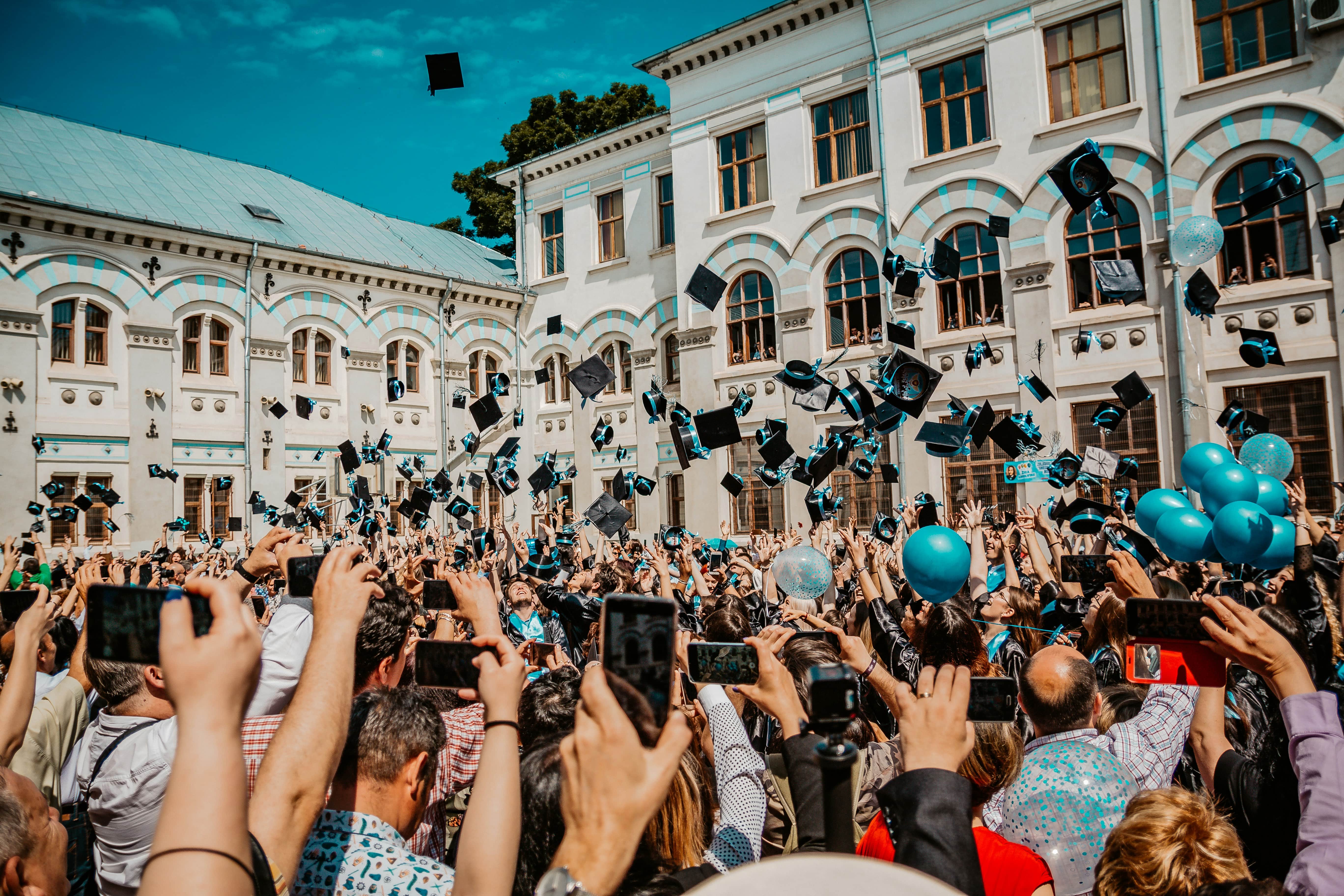 Crowd of graduates throwing black caps with blue ribbons into the air outside a historic building, many holding up phones and blue balloons.