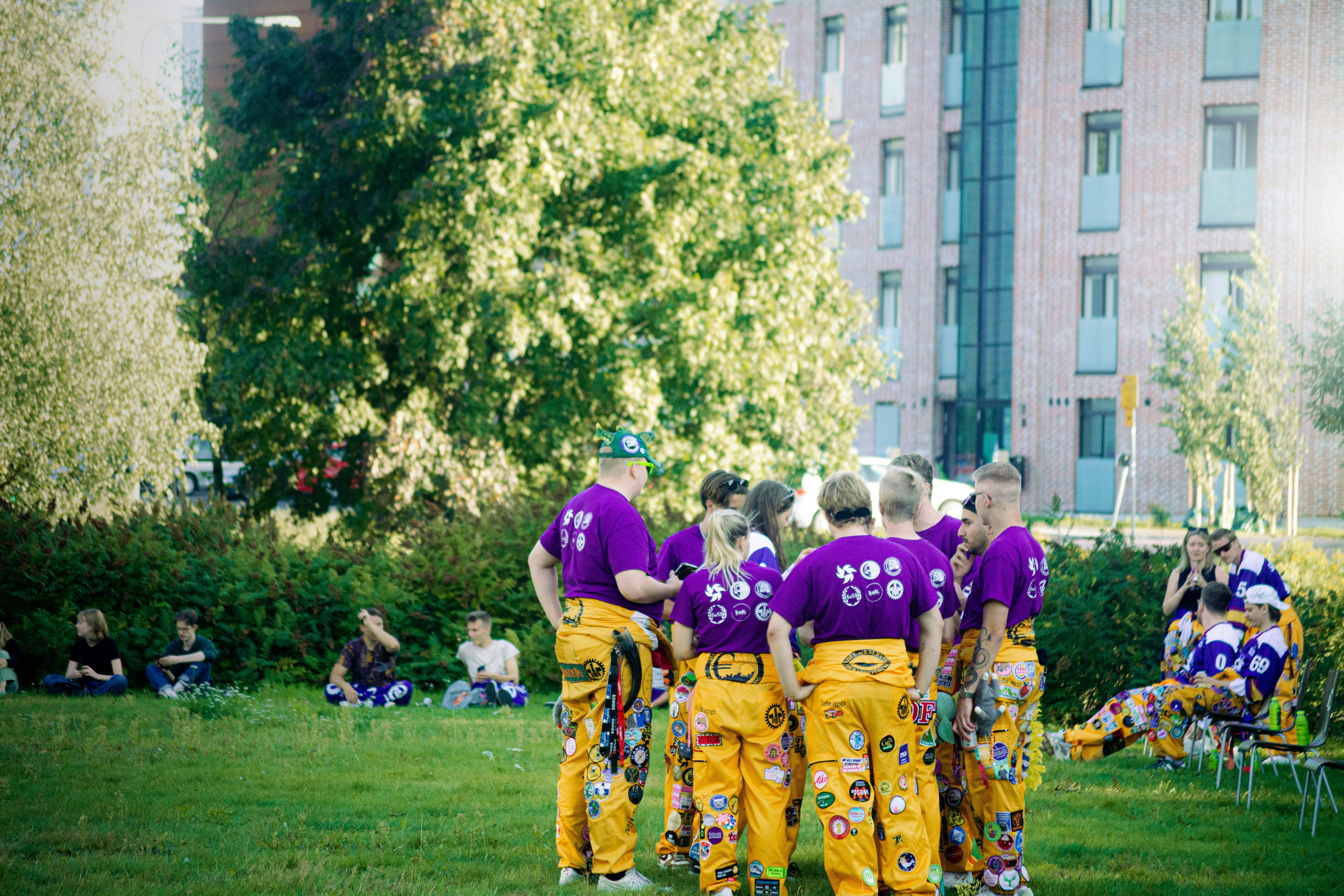 Group of people wearing matching purple shirts and yellow pants with patches gathered on grass near trees and a brick building.