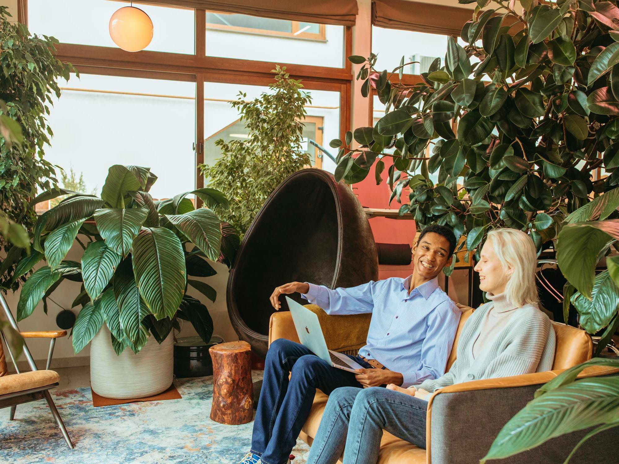 Two people sitting on a couch in a plant-filled room, one holding a laptop and smiling at the other.