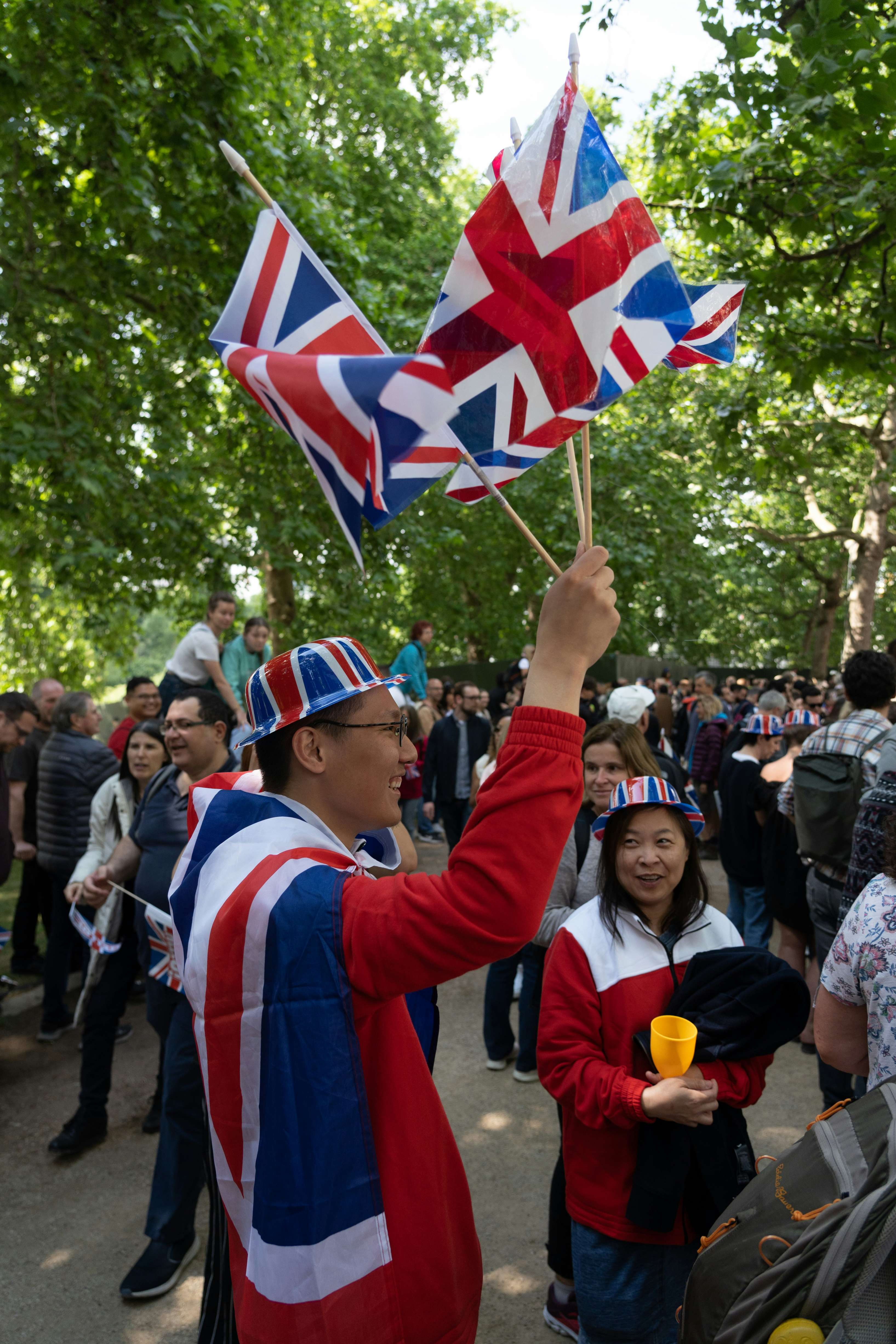 Crowd at an outdoor event with a man waving multiple Union Jack flags wearing a matching hat and cape.