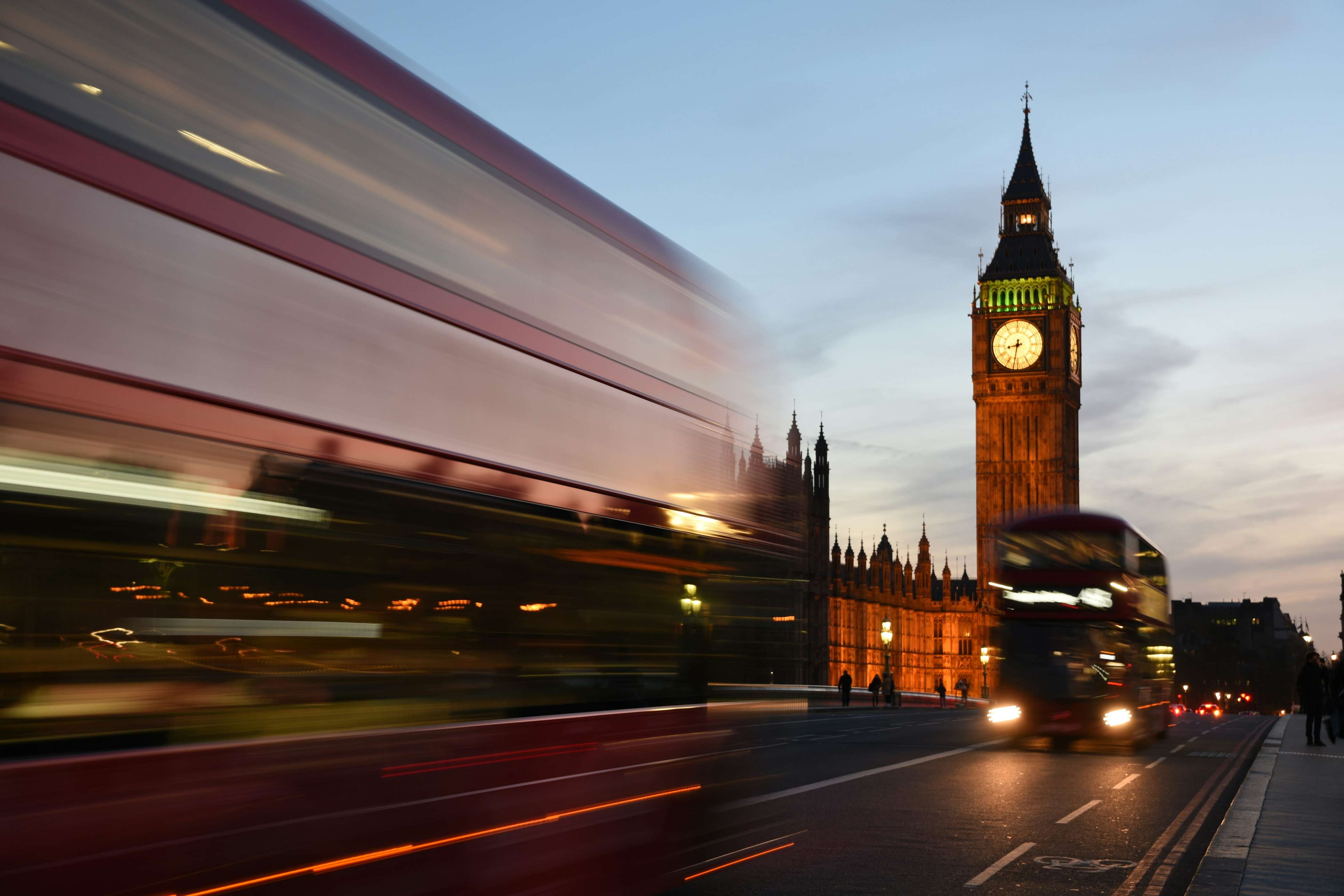 Blurred motion red double-decker buses passing near the illuminated Big Ben clock tower at dusk in London.
