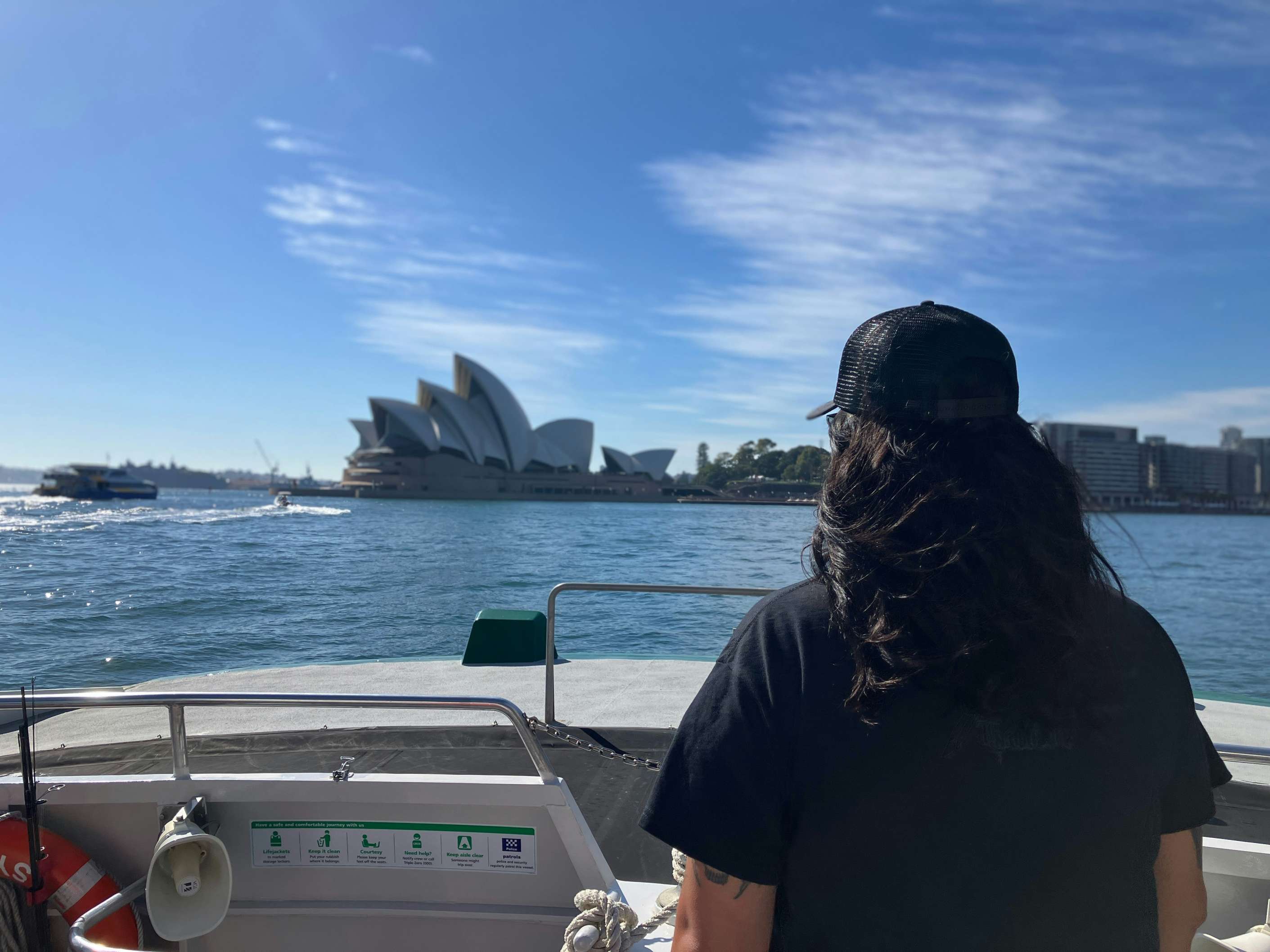 Person with long hair and a black cap standing on a boat facing Sydney Opera House across the water on a sunny day.