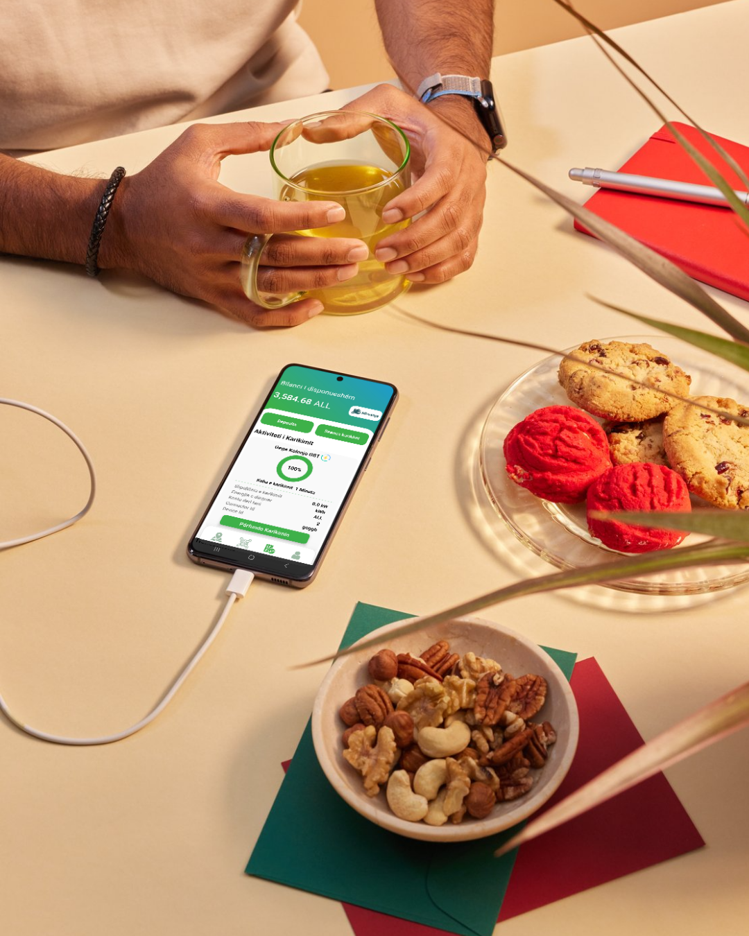 Person holding a glass cup of tea at a table with a smartphone displaying a green-yellow app screen, a plate of cookies, and a bowl of mixed nuts.