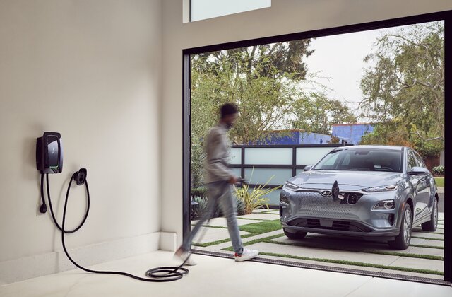 Person walking past a gray electric car parked outside a garage with a wall-mounted EV charger inside.