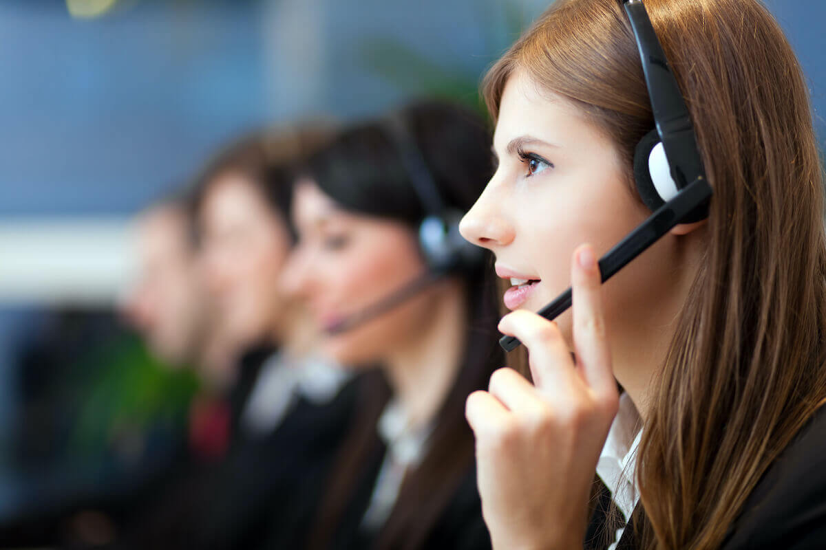 Close-up of a woman wearing a headset and speaking, with a blurred row of call center agents in the background.