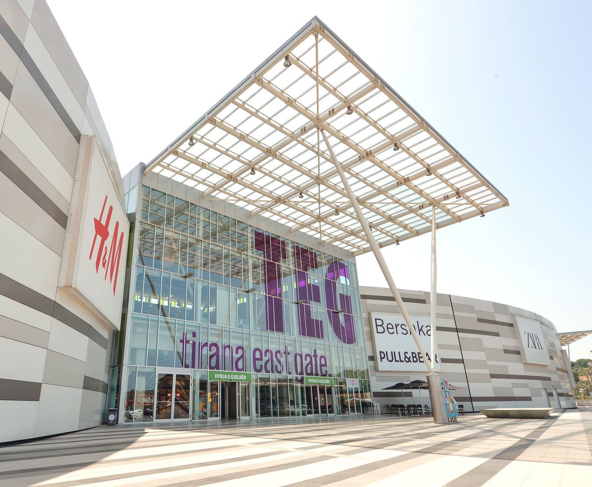 Entrance of Tirana East Gate shopping mall with large glass facade and signs for H&M, Bershka, Pull&Bear, and Zara stores.