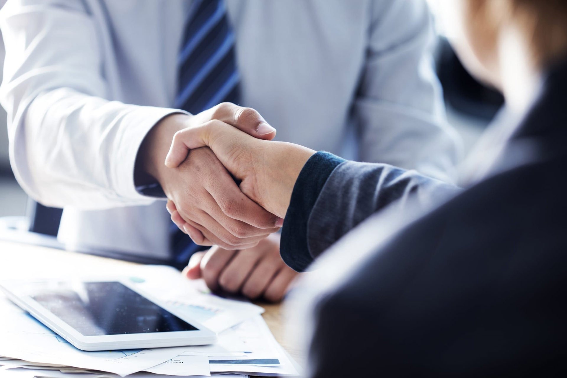 Two businesspeople shaking hands over a desk with documents and a tablet.