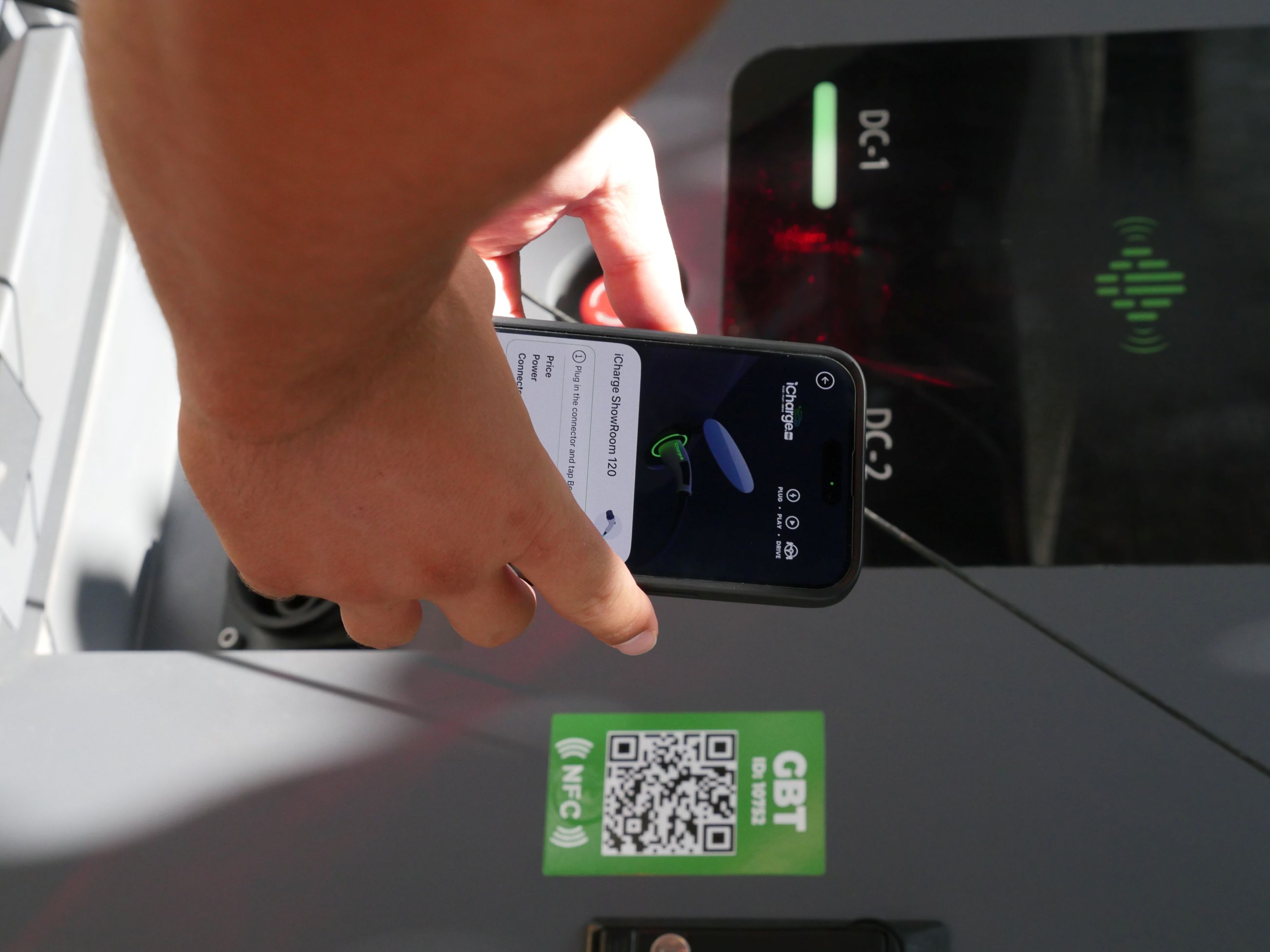 Person holding a smartphone displaying the iCharge ShowRoom 120 app near an electric vehicle charging station with a visible NFC label and QR code.