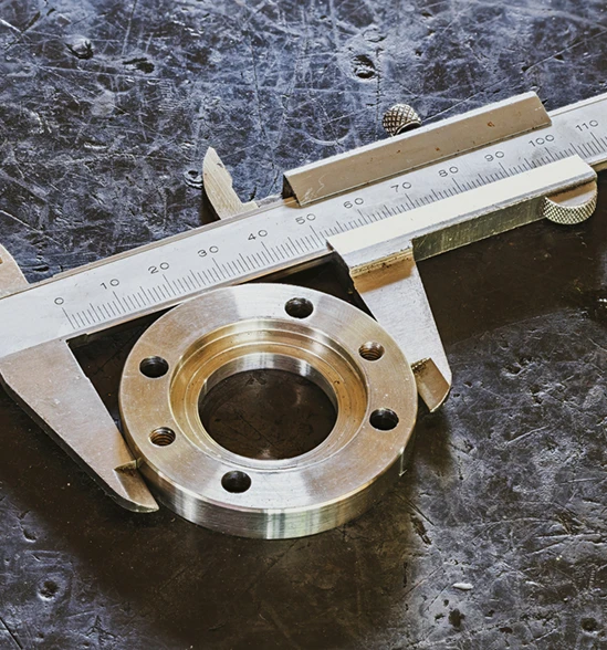 Metal caliper measuring diameter of a circular machined metal part with multiple holes on a scratched dark surface.