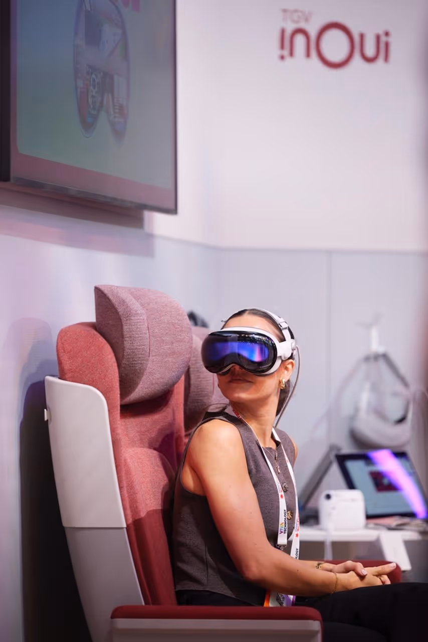 Woman seated in a pink cushioned chair wearing a VR headset with reflective lenses, looking slightly upward and to the side.