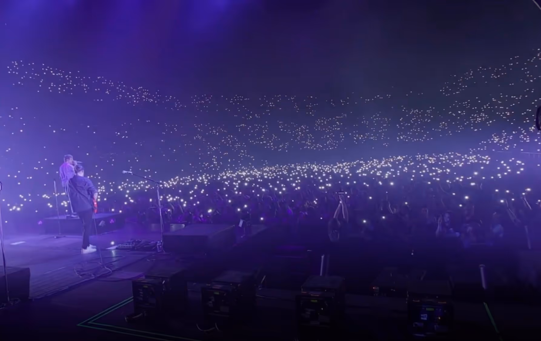 View from stage of a concert with two performers and a large audience holding up lights in a dark venue.
