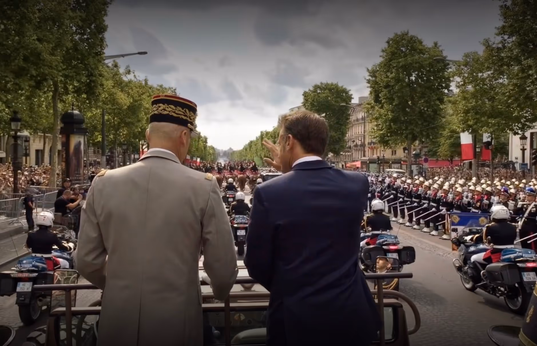 Two men, one in a military uniform and the other in a suit, standing in an open vehicle during a formal parade with marching soldiers and motorcyclists lined up along a tree-lined street filled with spectators.