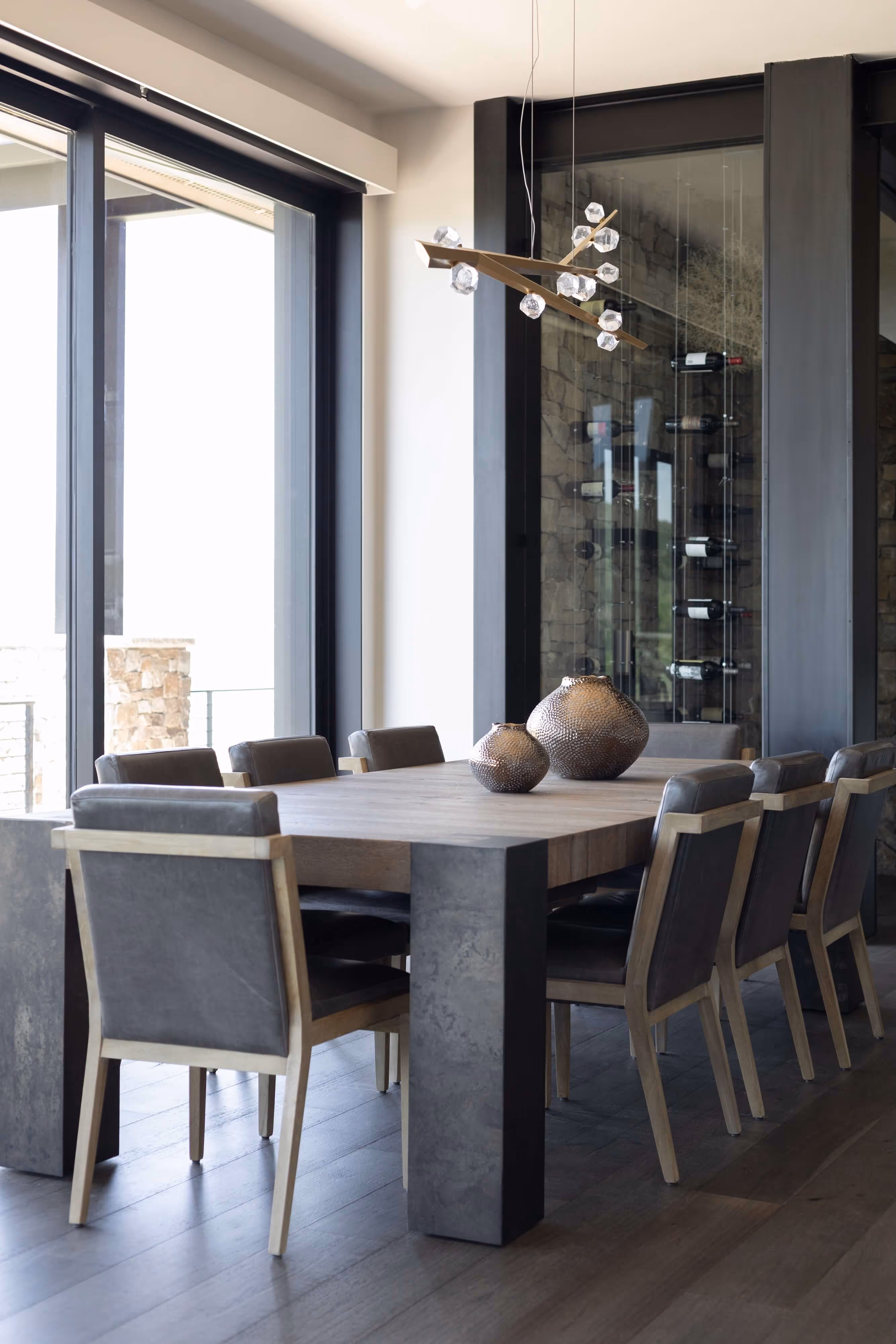 Modern dining room with large wooden table, eight gray cushioned chairs, decorative vases, and floor-to-ceiling windows.