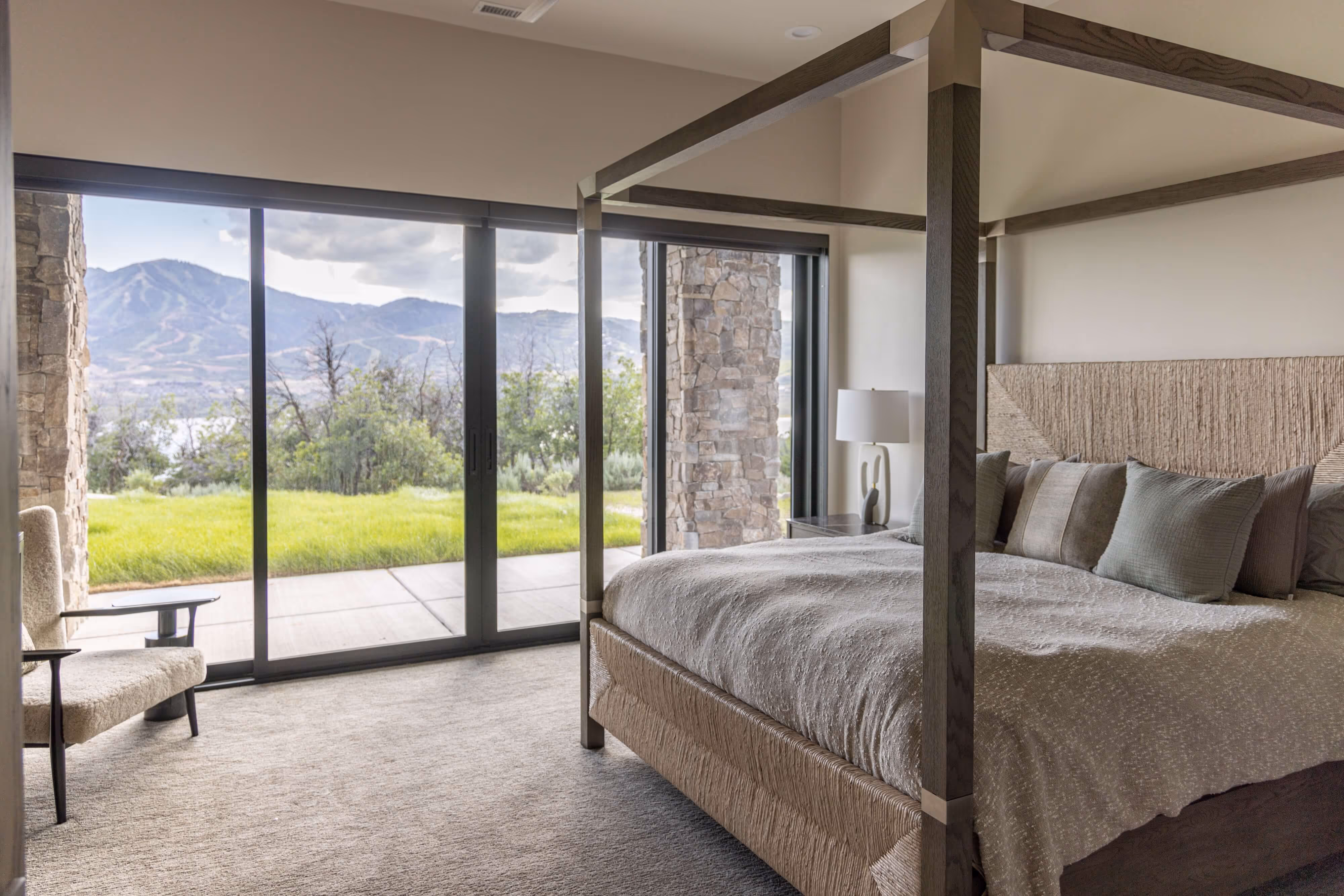 Modern bedroom with a canopy bed, neutral bedding, and large sliding glass doors showing a mountain and grassy outdoor view.