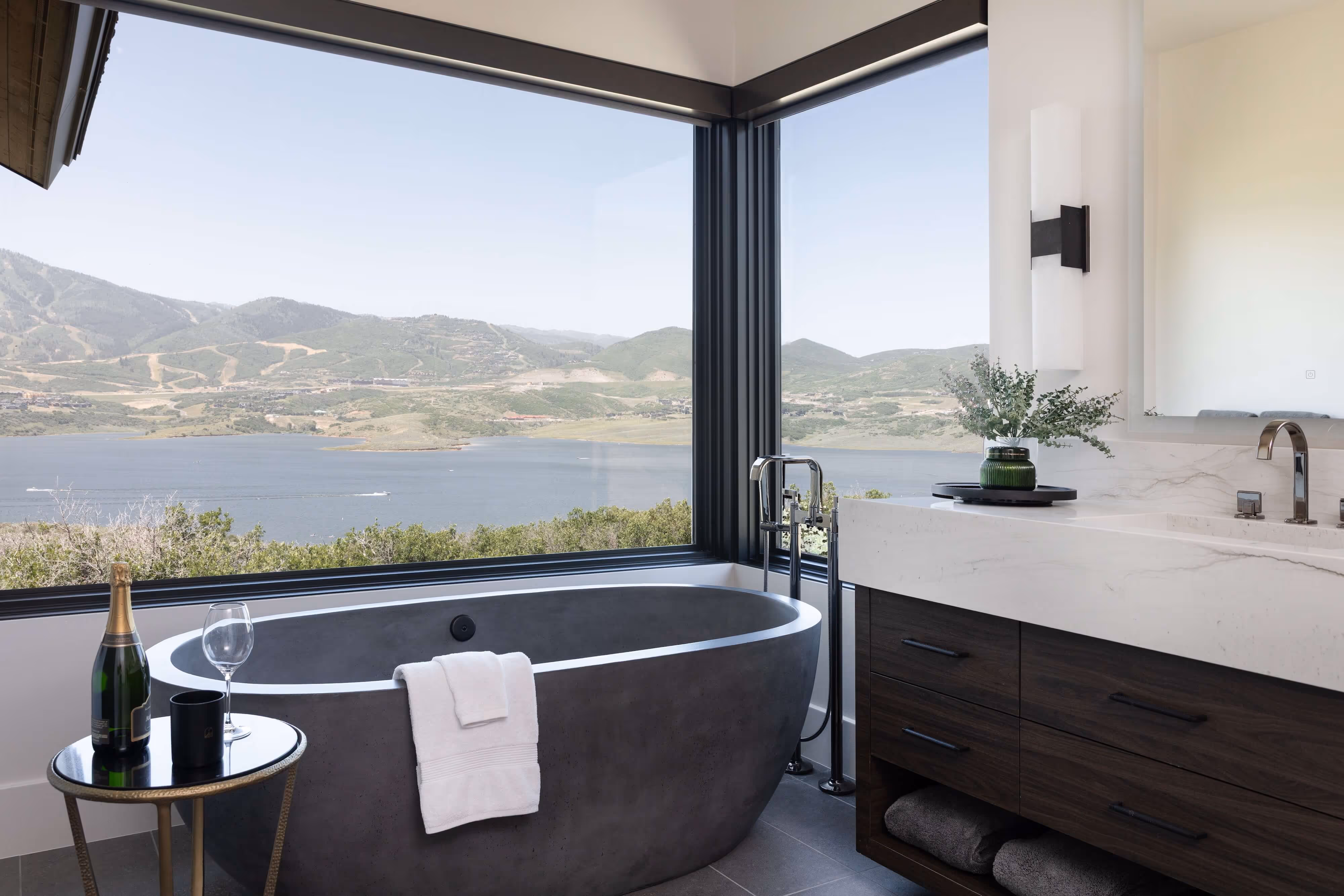 Modern bathroom with dark freestanding bathtub facing large corner windows overlooking a lake and mountains, next to vanity with sink and a small table holding a champagne bottle and glass.