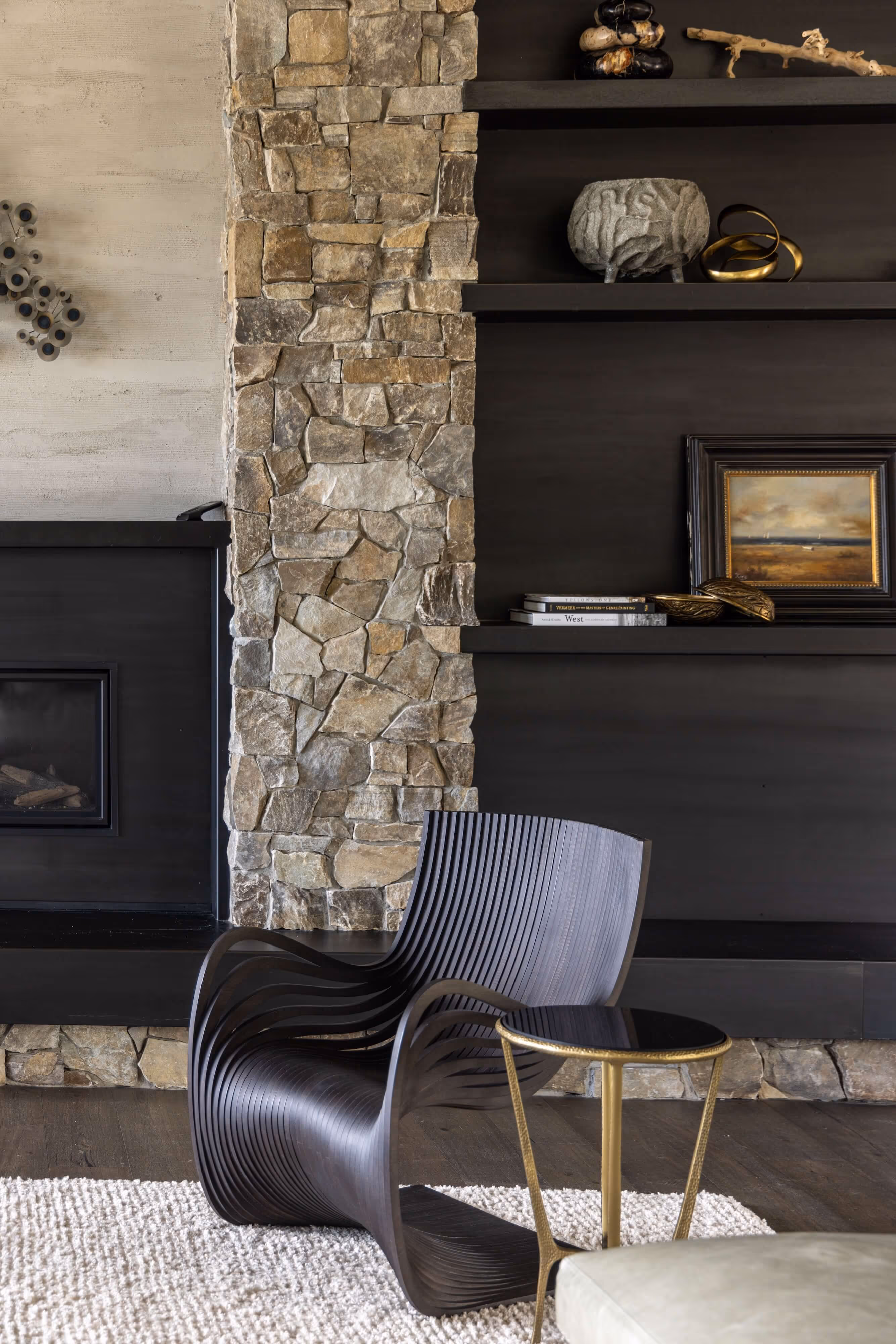 Modern dark wood sculptural chair next to a small round black and gold side table in a living room with stone fireplace and dark shelves.