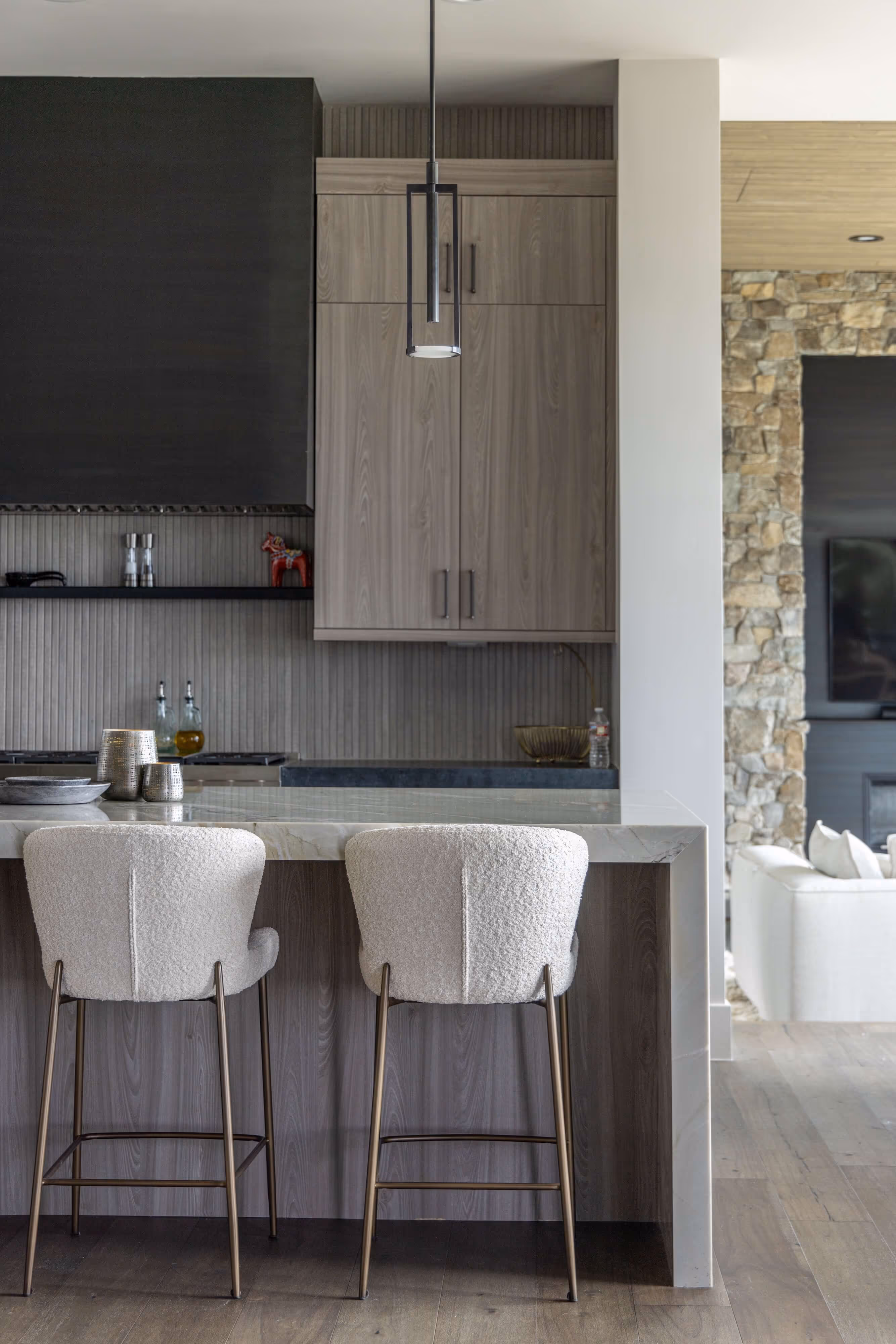 Modern kitchen with marble island, two white upholstered bar stools, wood cabinets, and pendant light above.