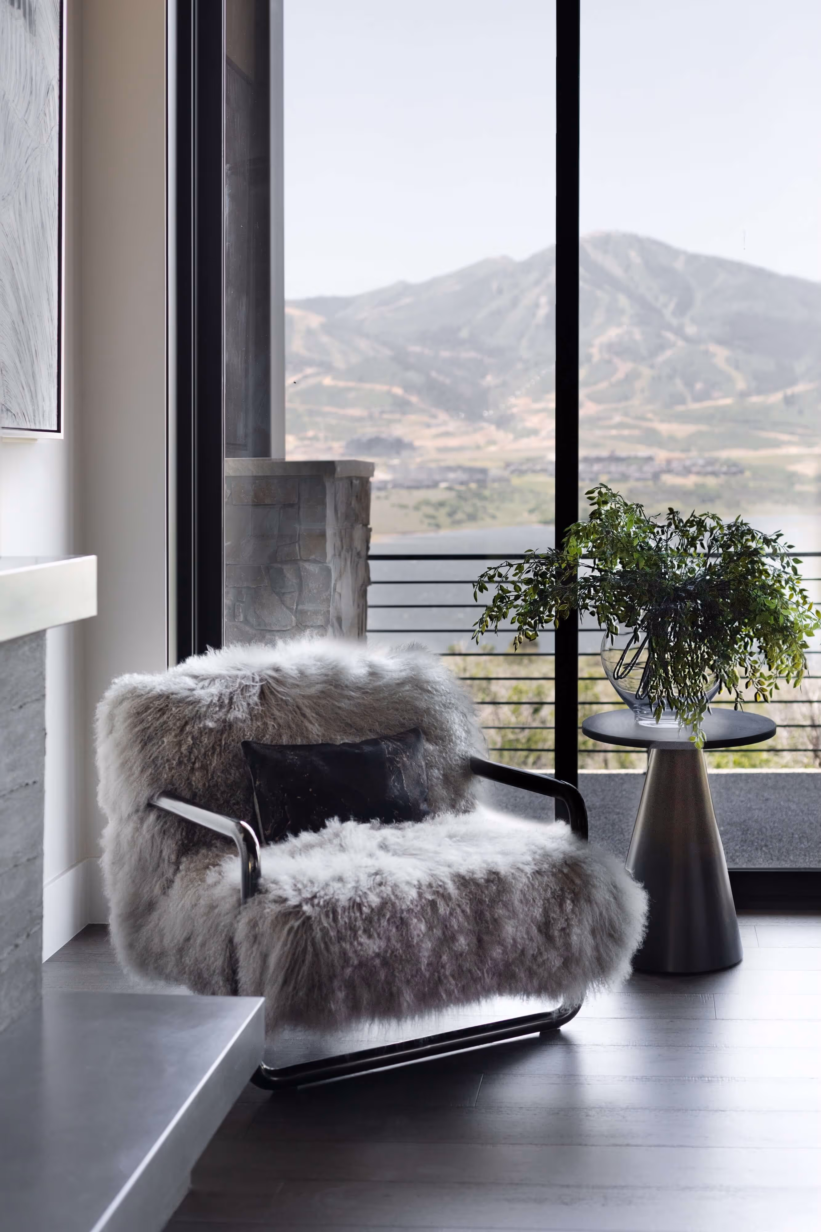 Cozy gray fluffy armchair with black pillow next to a round side table with a green plant, overlooking a mountain landscape through large glass doors.