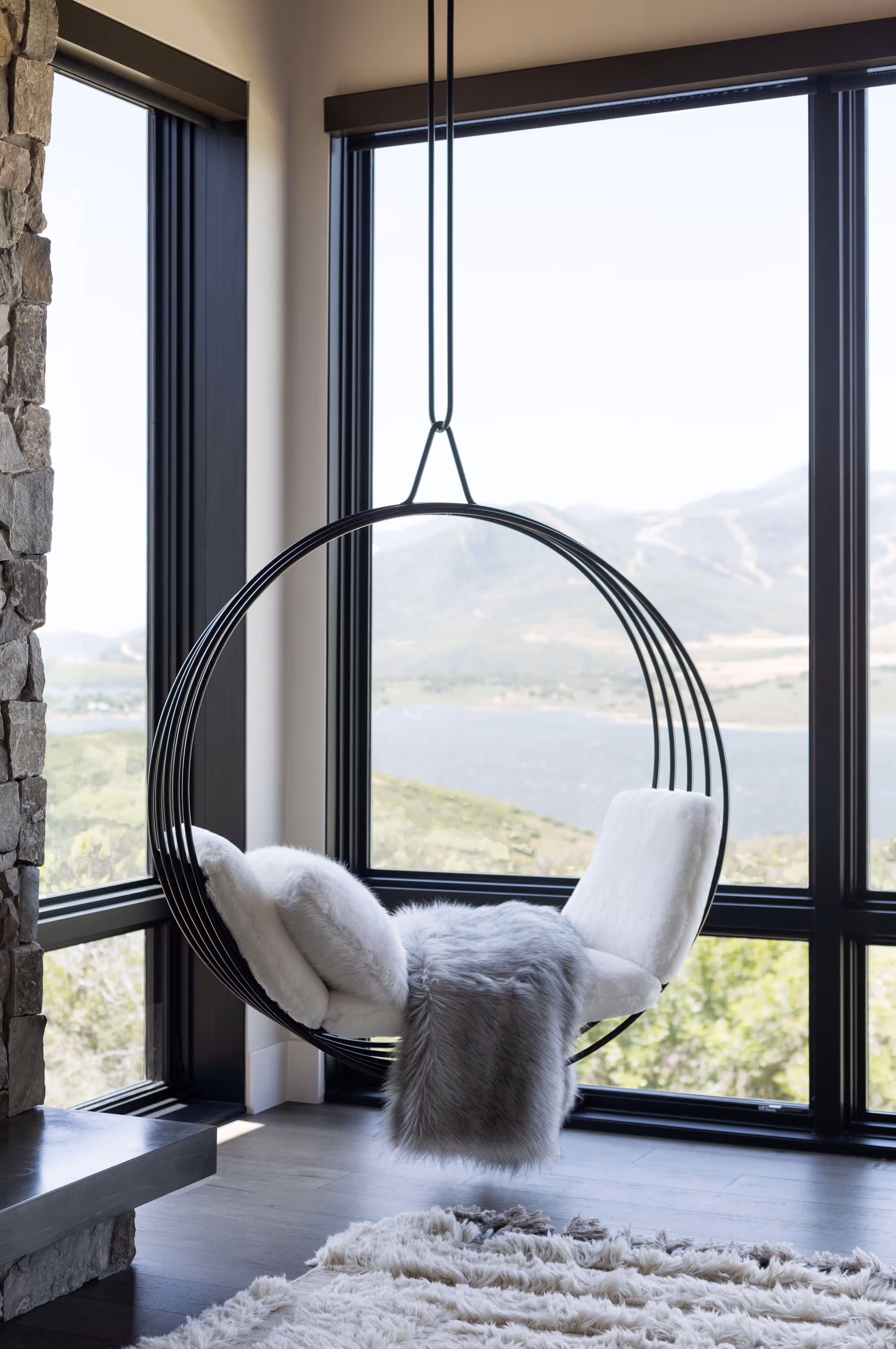 Modern black metal hanging chair with white cushions and gray fur throw by large windows overlooking mountains and lake.