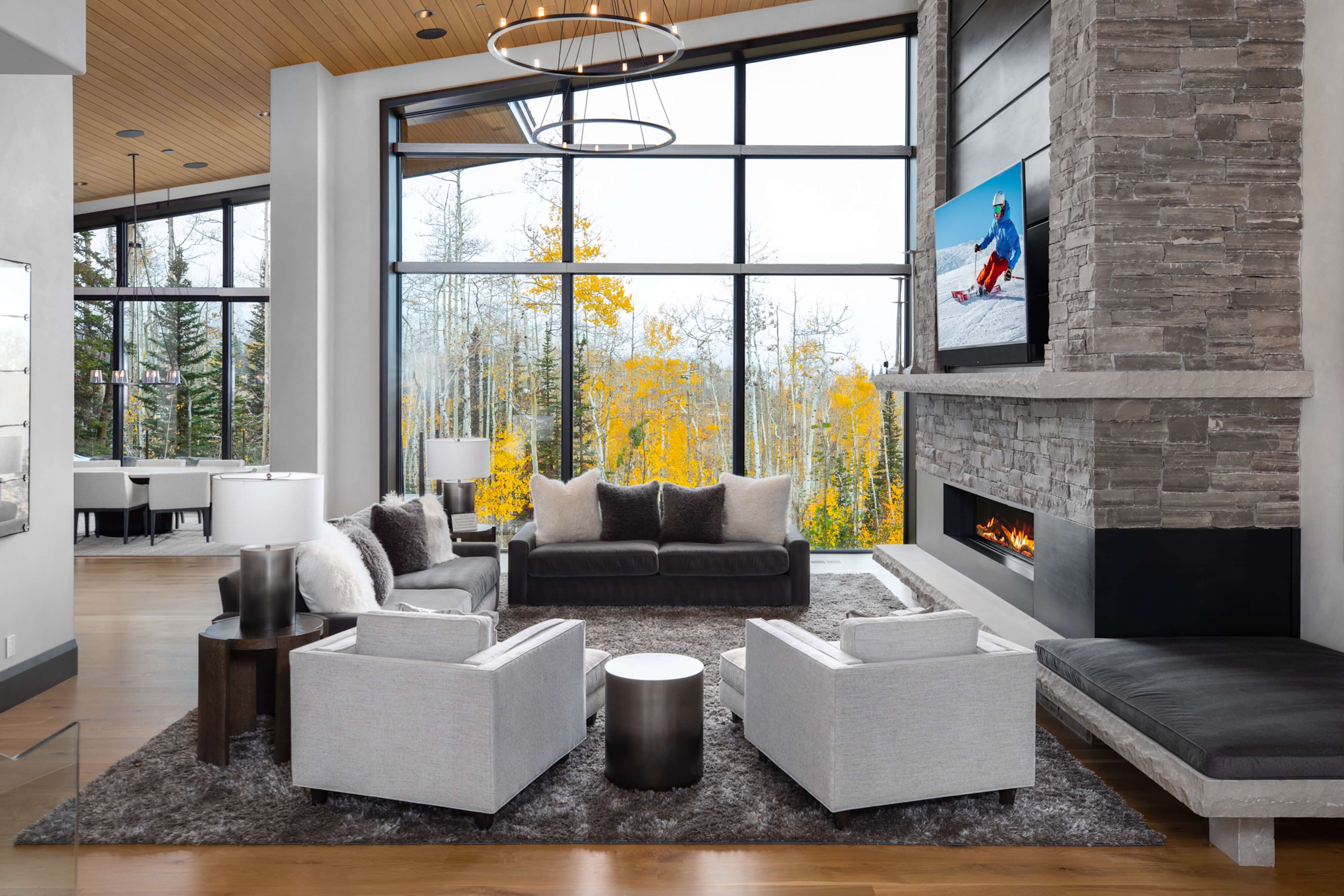 Modern living room with large floor-to-ceiling windows showing autumn trees, gray sofas and armchairs, stone fireplace with TV above, and wooden flooring.