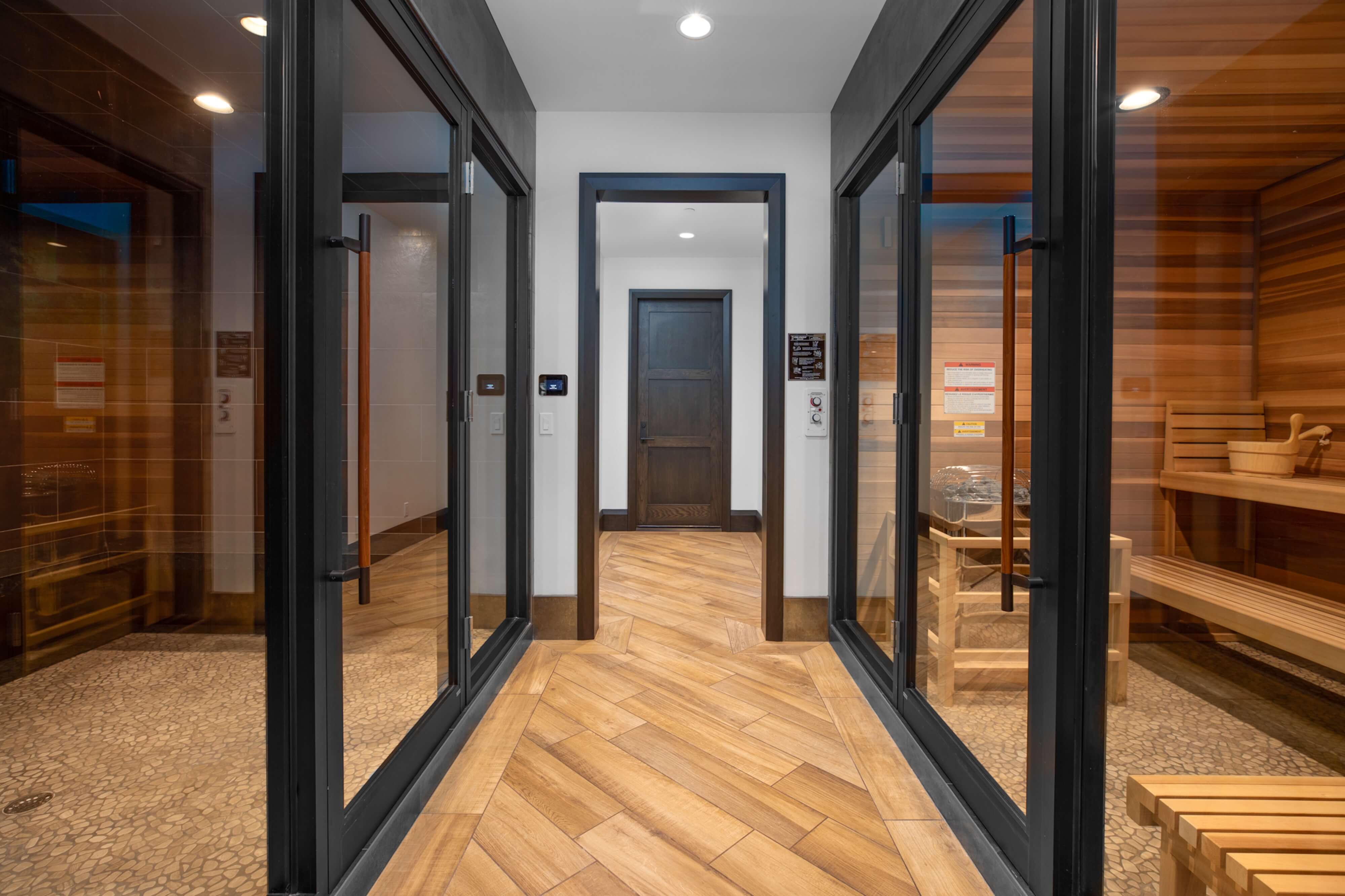 Corridor with wooden herringbone floor leading to a dark wooden door, flanked by glass doors revealing a steam room on the left and a sauna on the right.