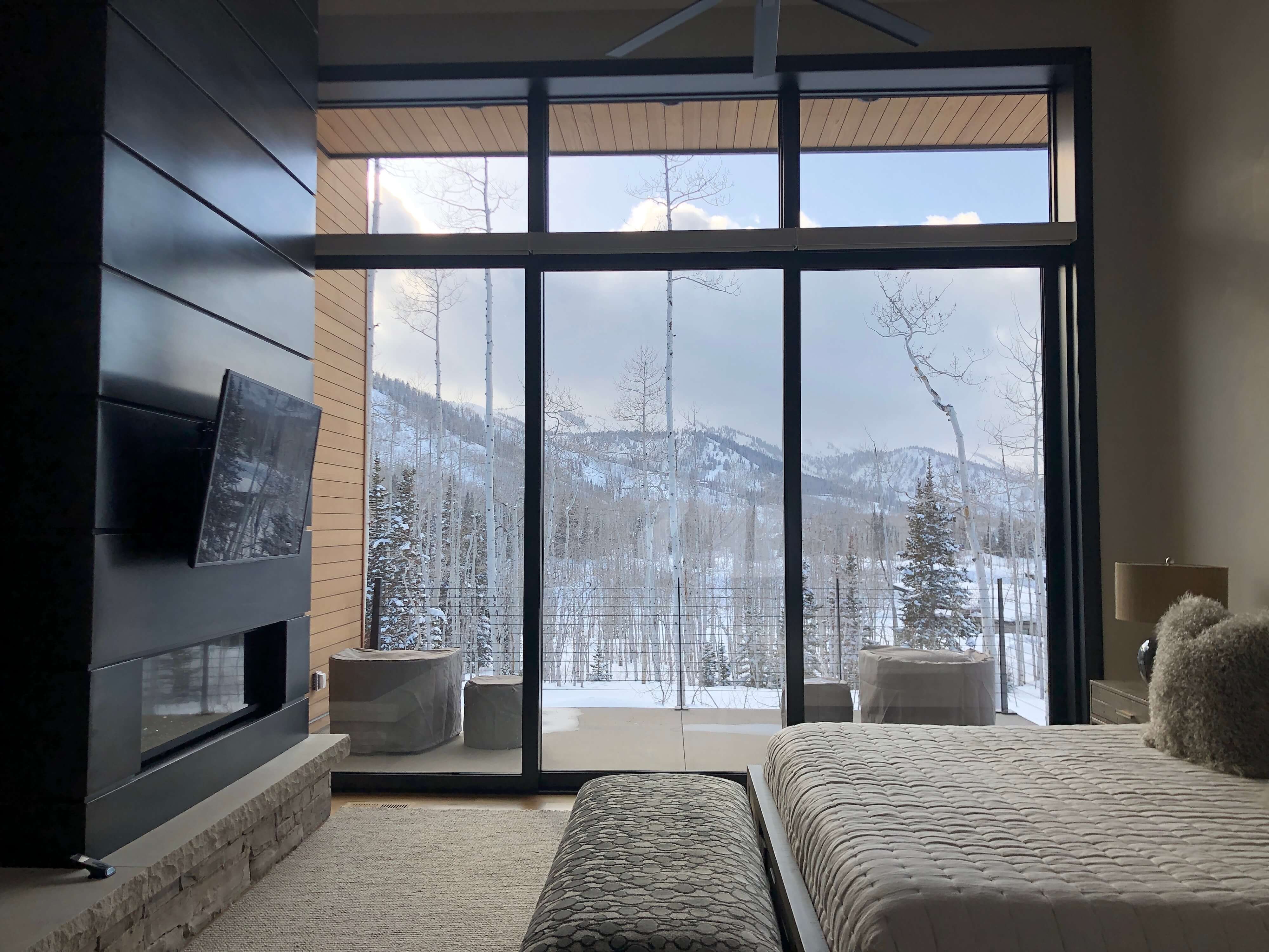 Modern bedroom with a large glass wall overlooking a snowy mountain landscape and bare trees.
