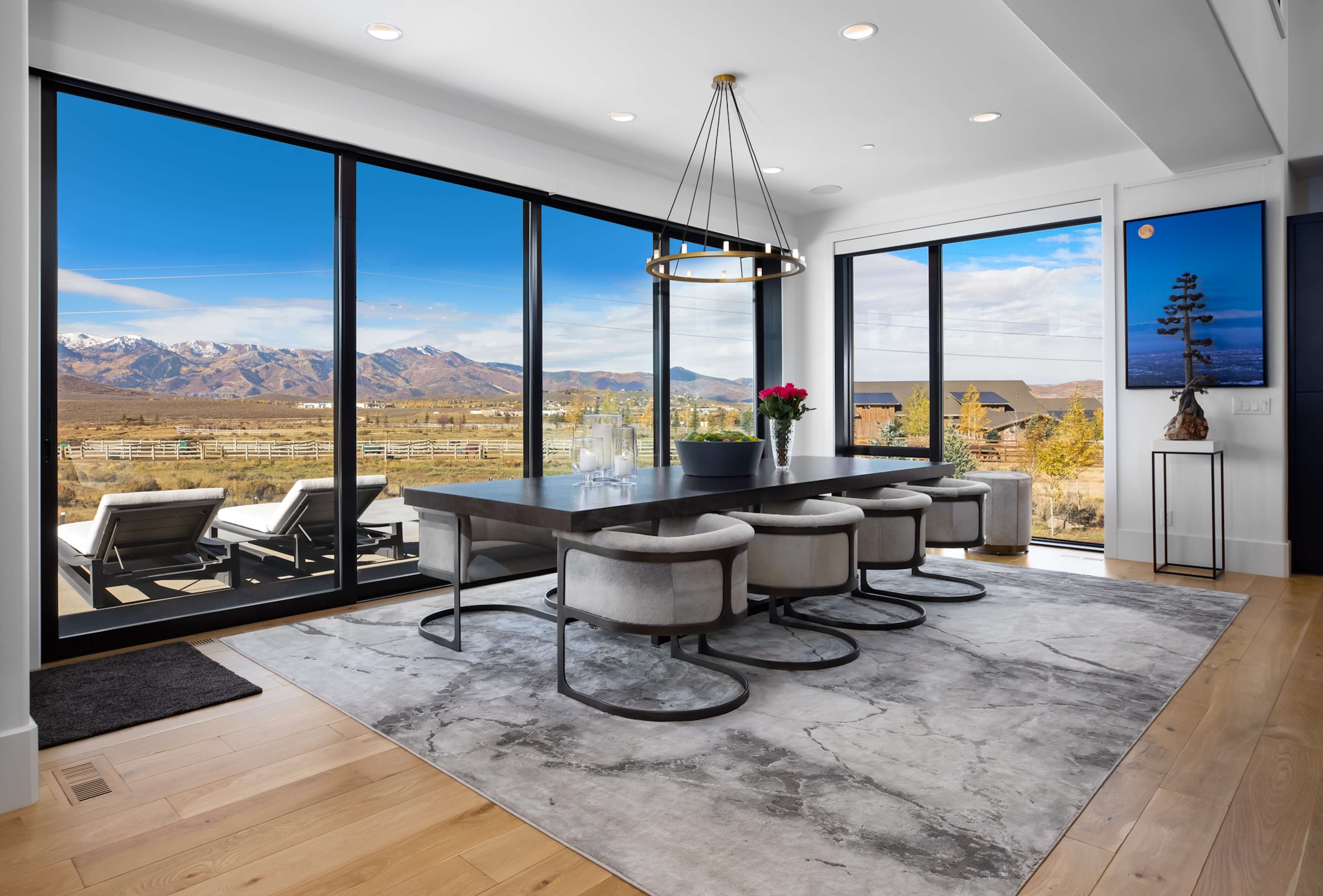 Modern dining room with a large dark wood table, six gray cushioned chairs, a circular chandelier, and floor-to-ceiling windows showing mountain and countryside views.