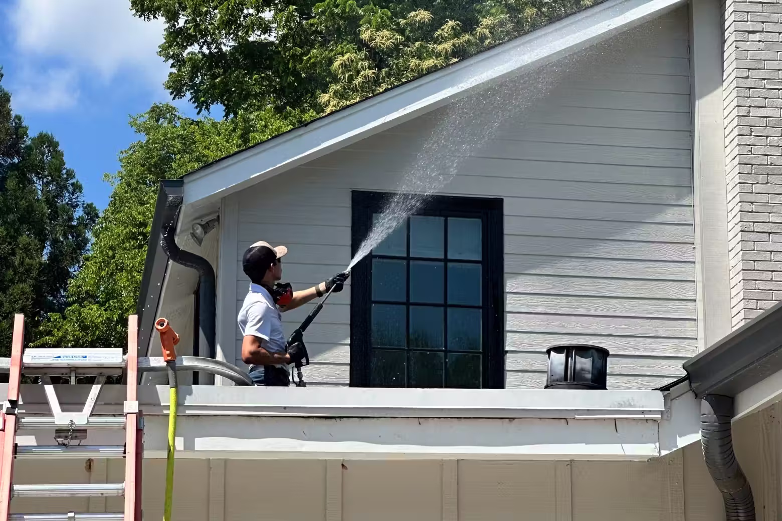 Man soft washing the siding of a house near a dark-paned window and gutter.