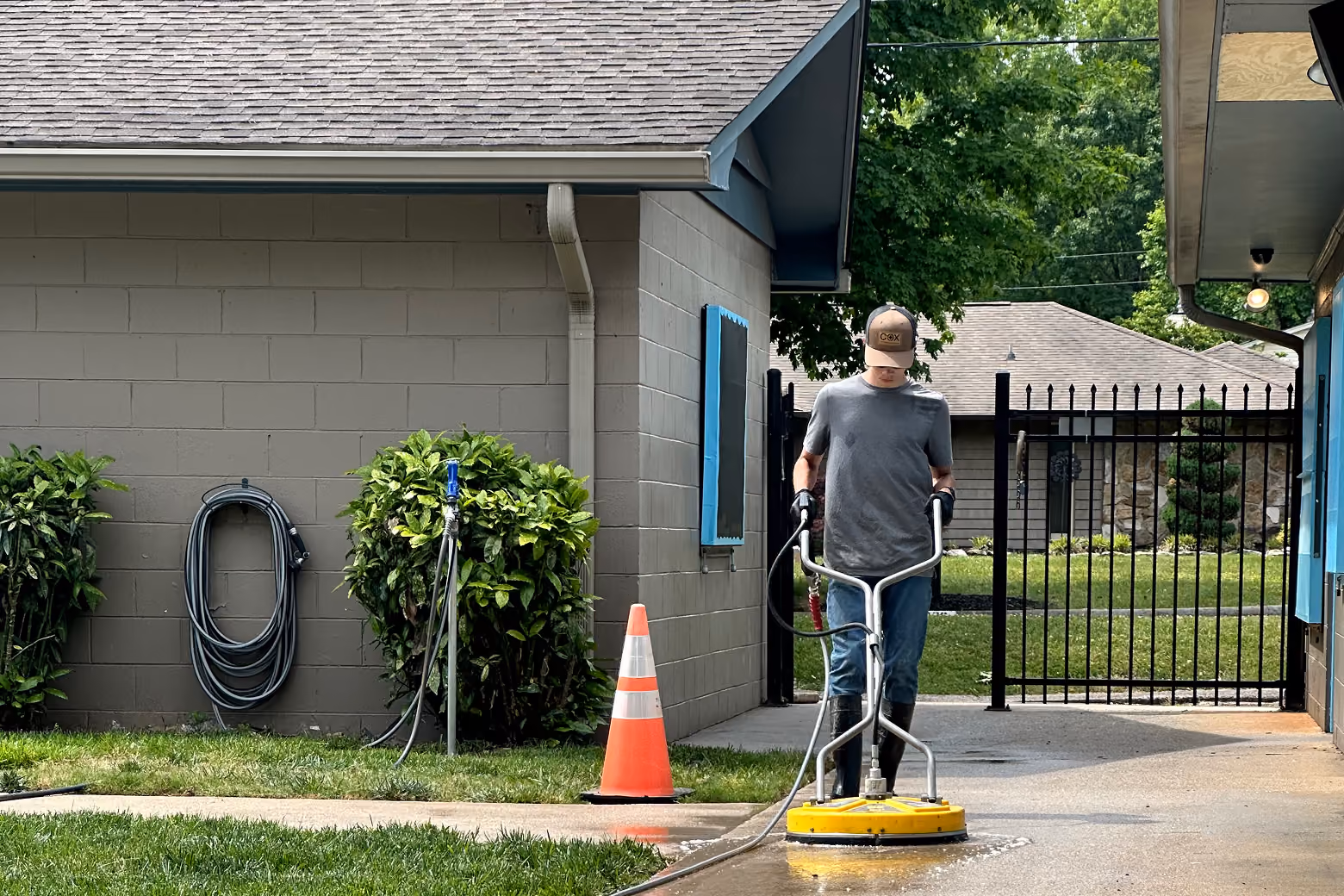 Person pressure washing a concrete walkway in front of a grey building with bushes and a fence.