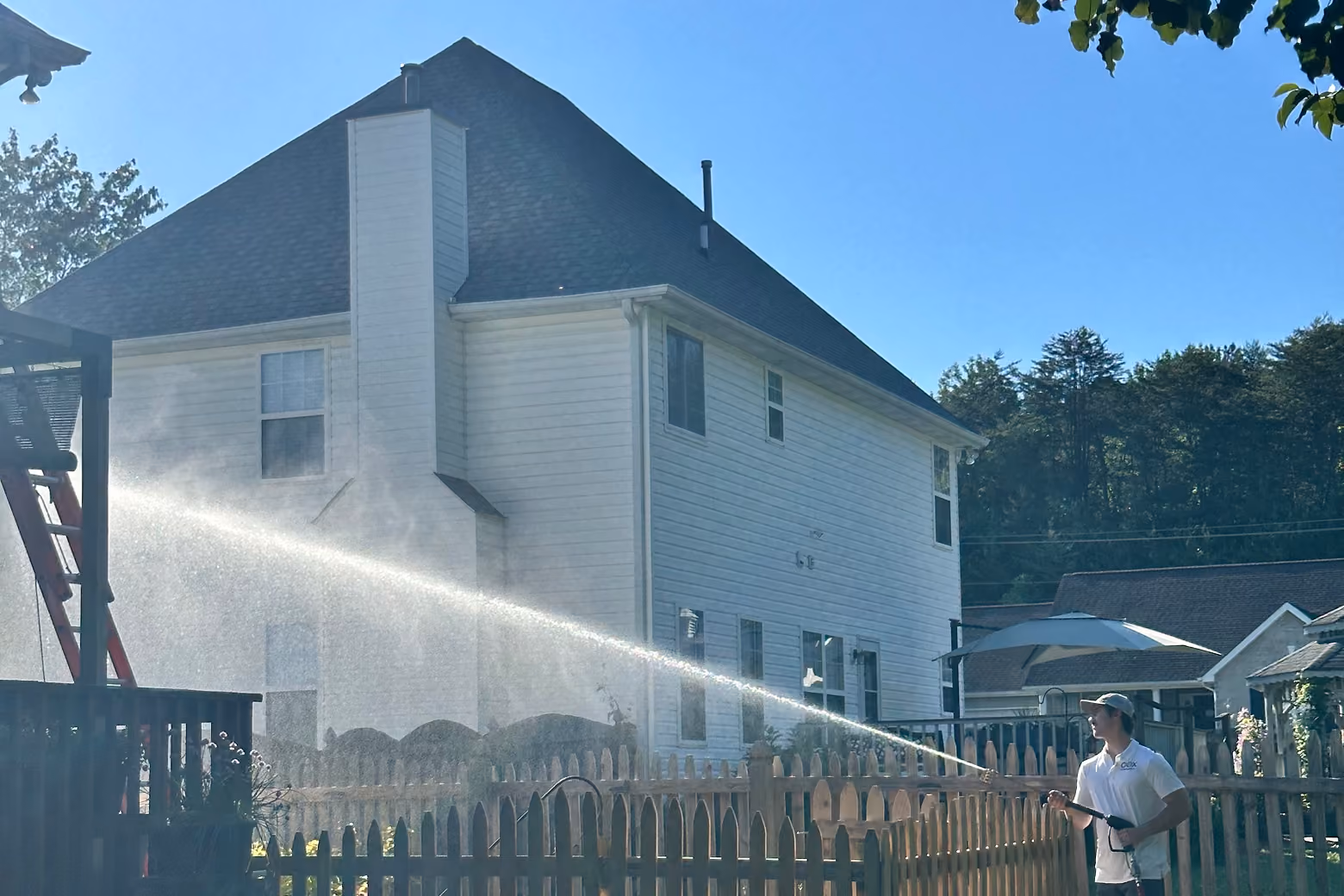 A man in a white shirt and hat pressure washes the side of a white house with a large chimney.