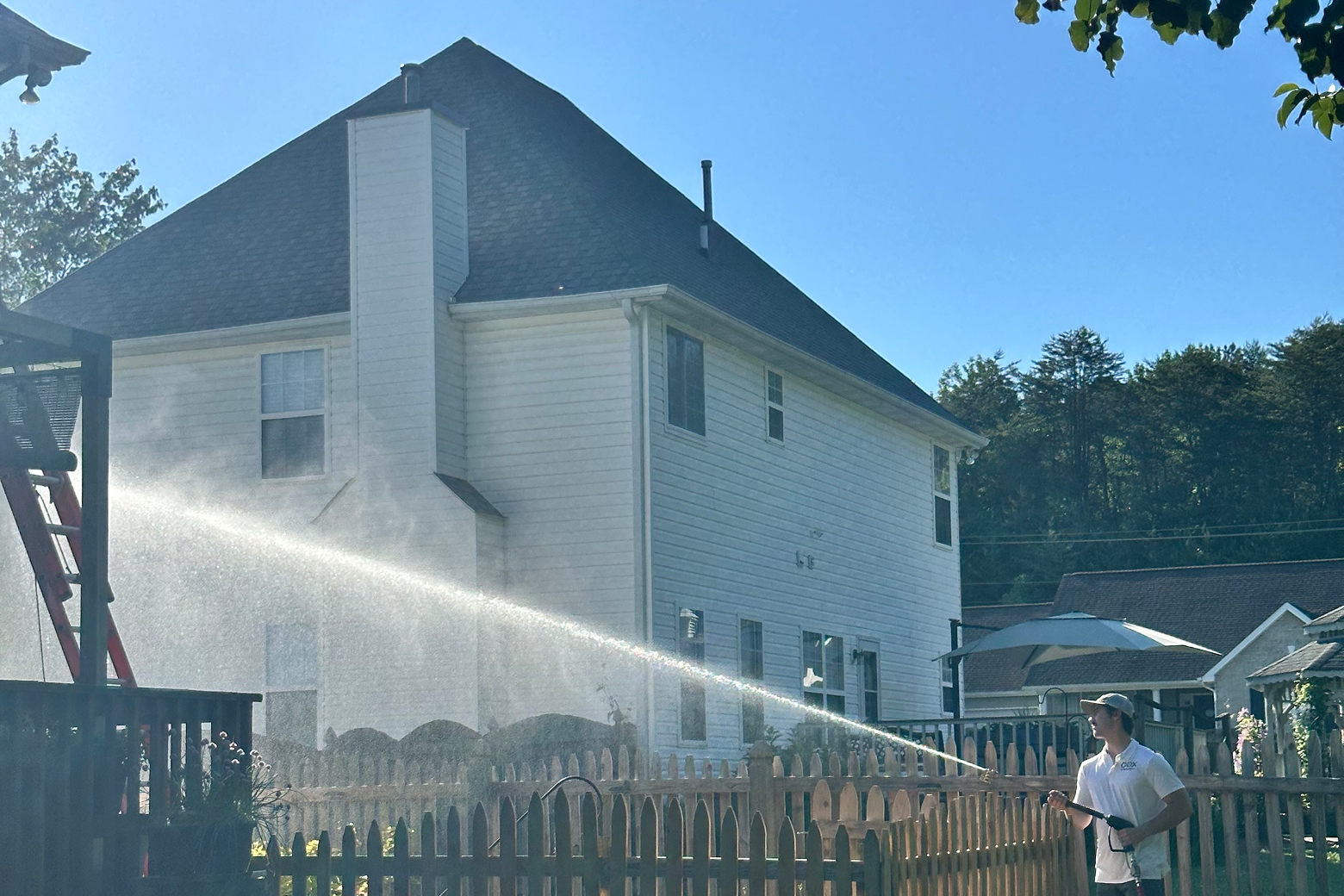 A man in a white shirt and hat pressure washes the side of a white house with a large chimney.