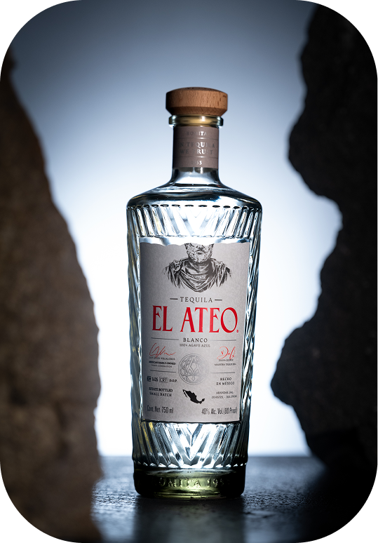 Bottle of El Ateo Blanco Tequila with a wooden cap on a dark surface between two rocks.