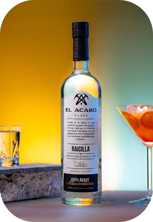 Bottle of El Acabo Plata Raicilla, a 100% agave spirit, next to a cocktail and a glass on a bar counter.