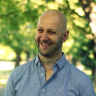 Smiling bald man with a beard wearing a blue checkered shirt standing outdoors with trees in the blurred background.