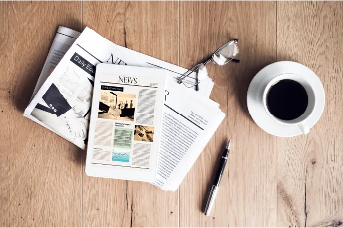 Newspapers, a pair of eyeglasses, a pen, and a cup of coffee on a wooden table.