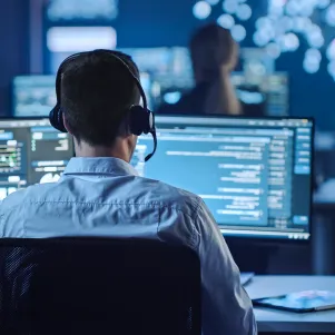 Person wearing headset working on multiple computer monitors displaying code in a dimly lit office.